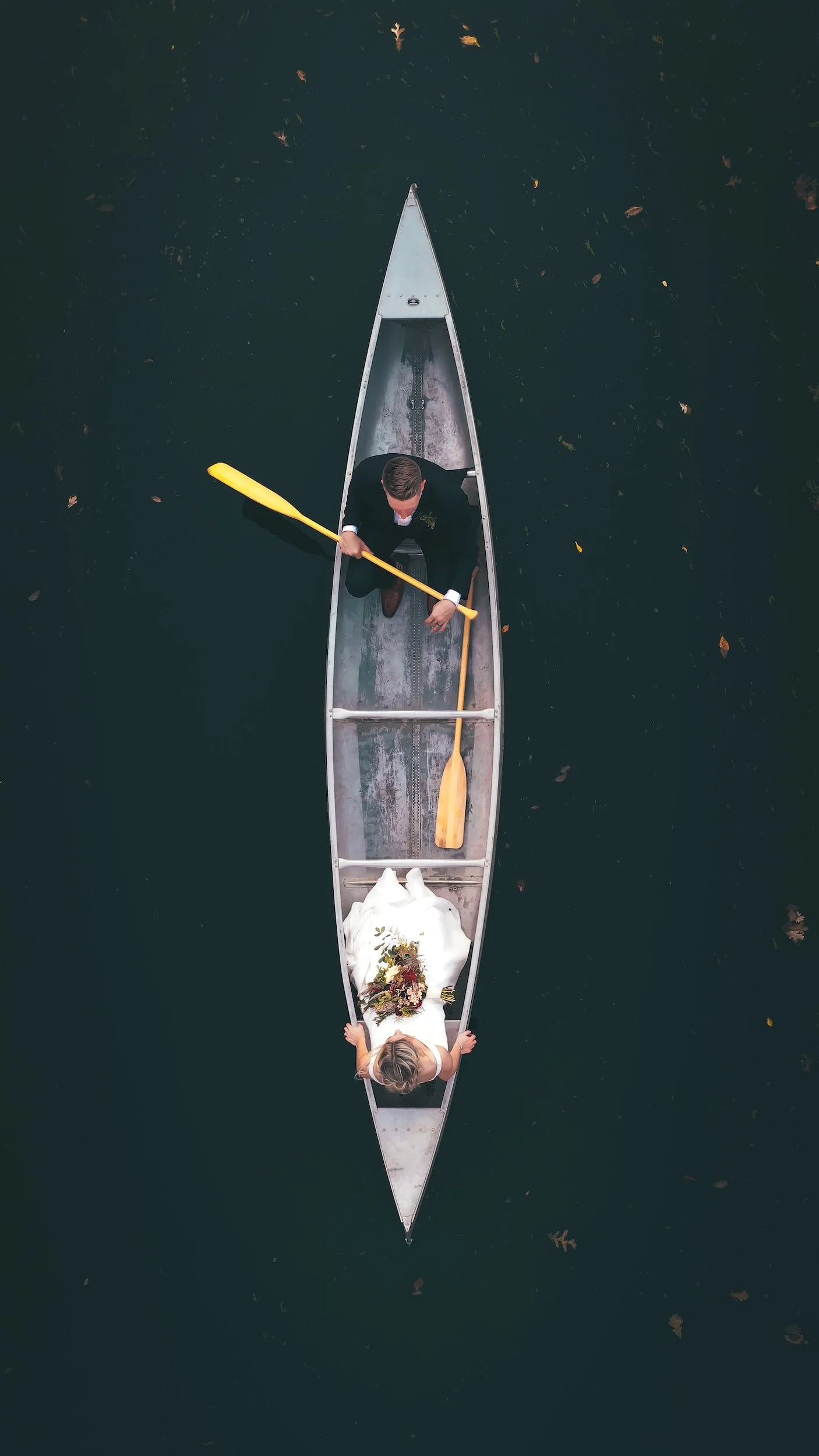 A woman in a wedding dress sitting in a boat holding a bouquet, with a man in a suit paddling the boat on water with leaf debris.