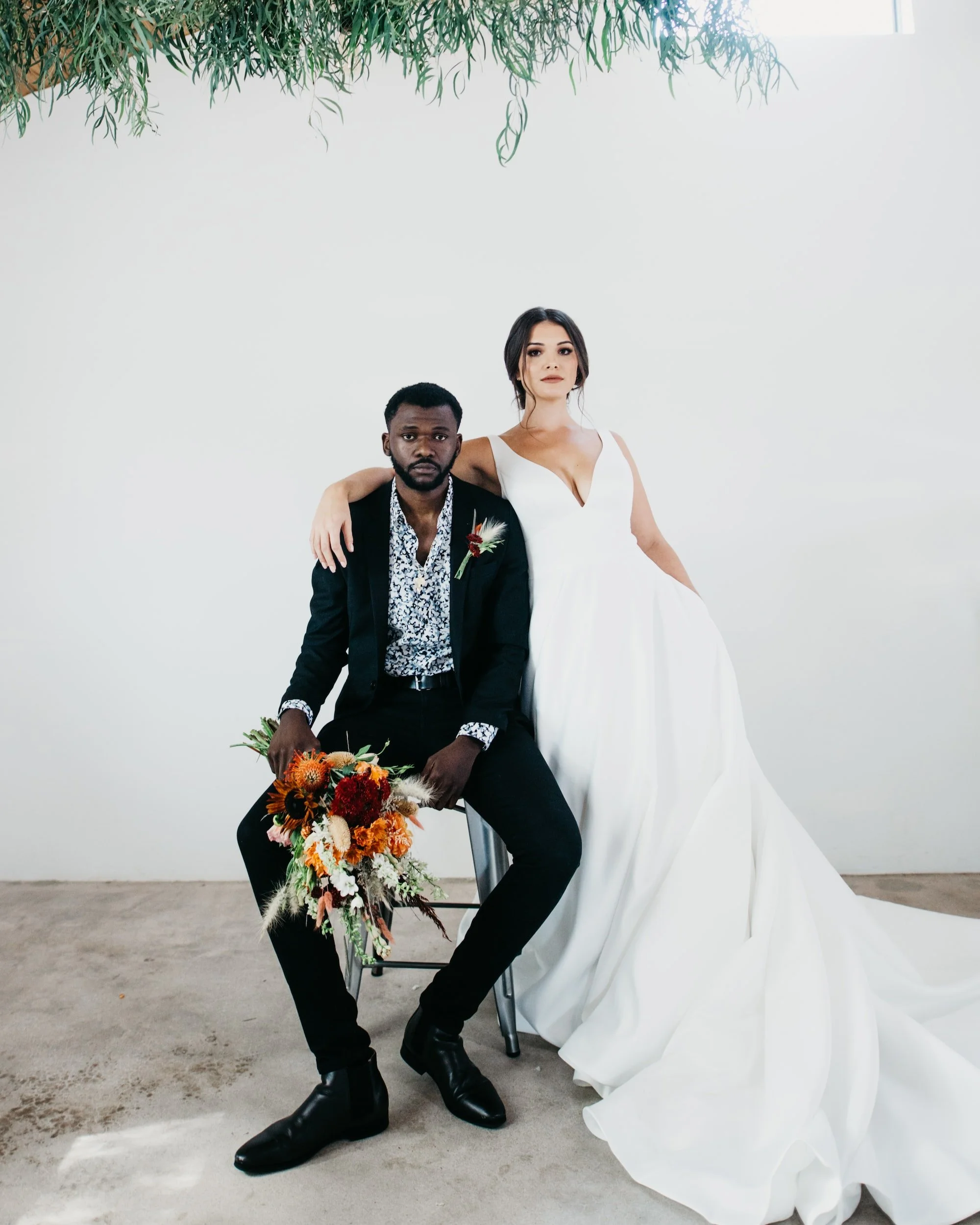 A couple on their wedding day, with the woman in a white gown and the man in a black suit, posing against a white wall with a green plant hanging above.