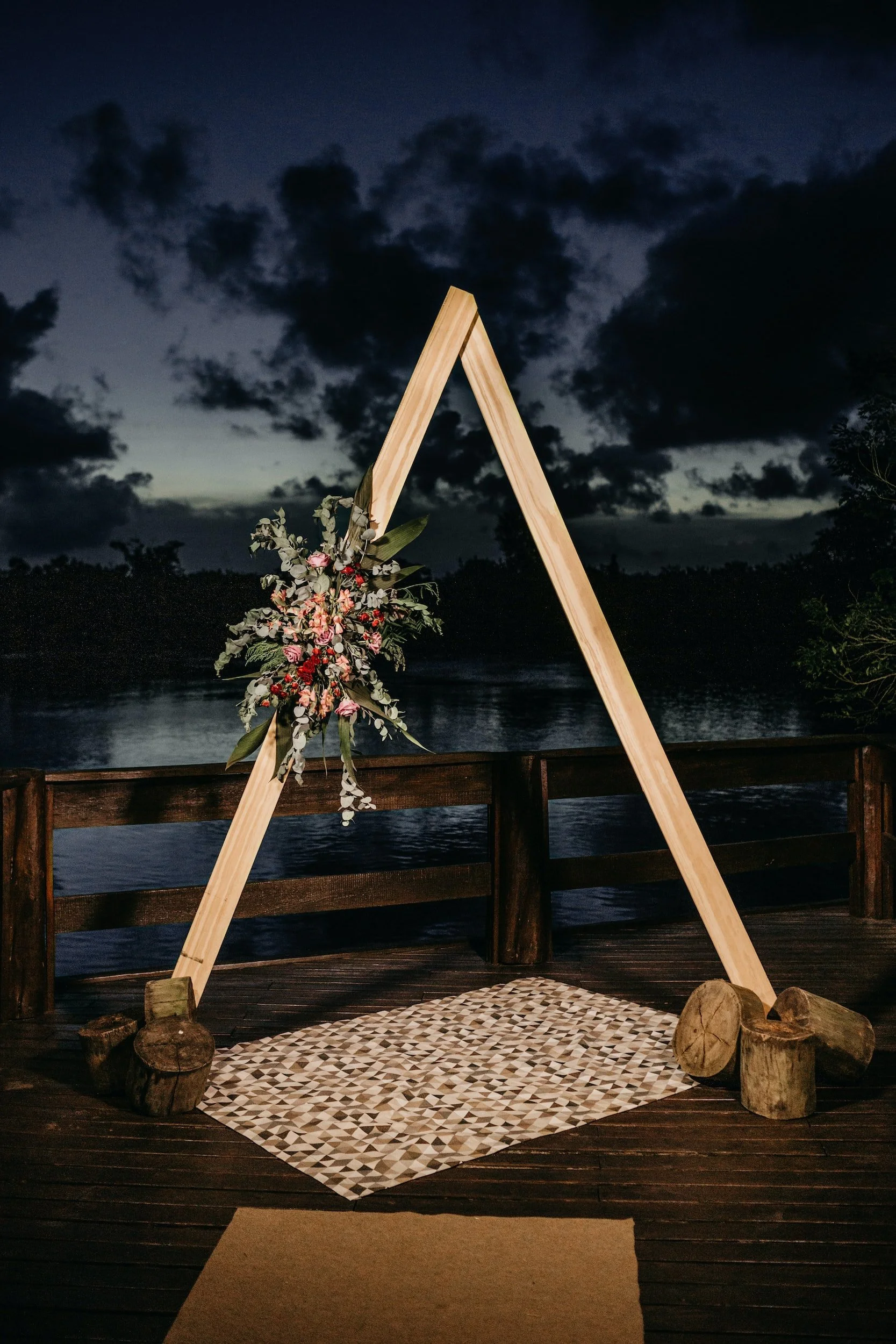 Wooden wedding arch decorated with flowers on a deck overlooking a river at dusk.