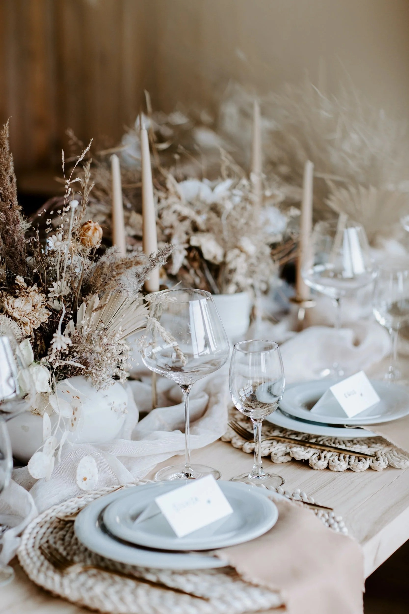 Elegant table setting with white plates, clear wine glasses, cutlery, and a centerpiece of dried flowers and candles on a beige tablecloth.