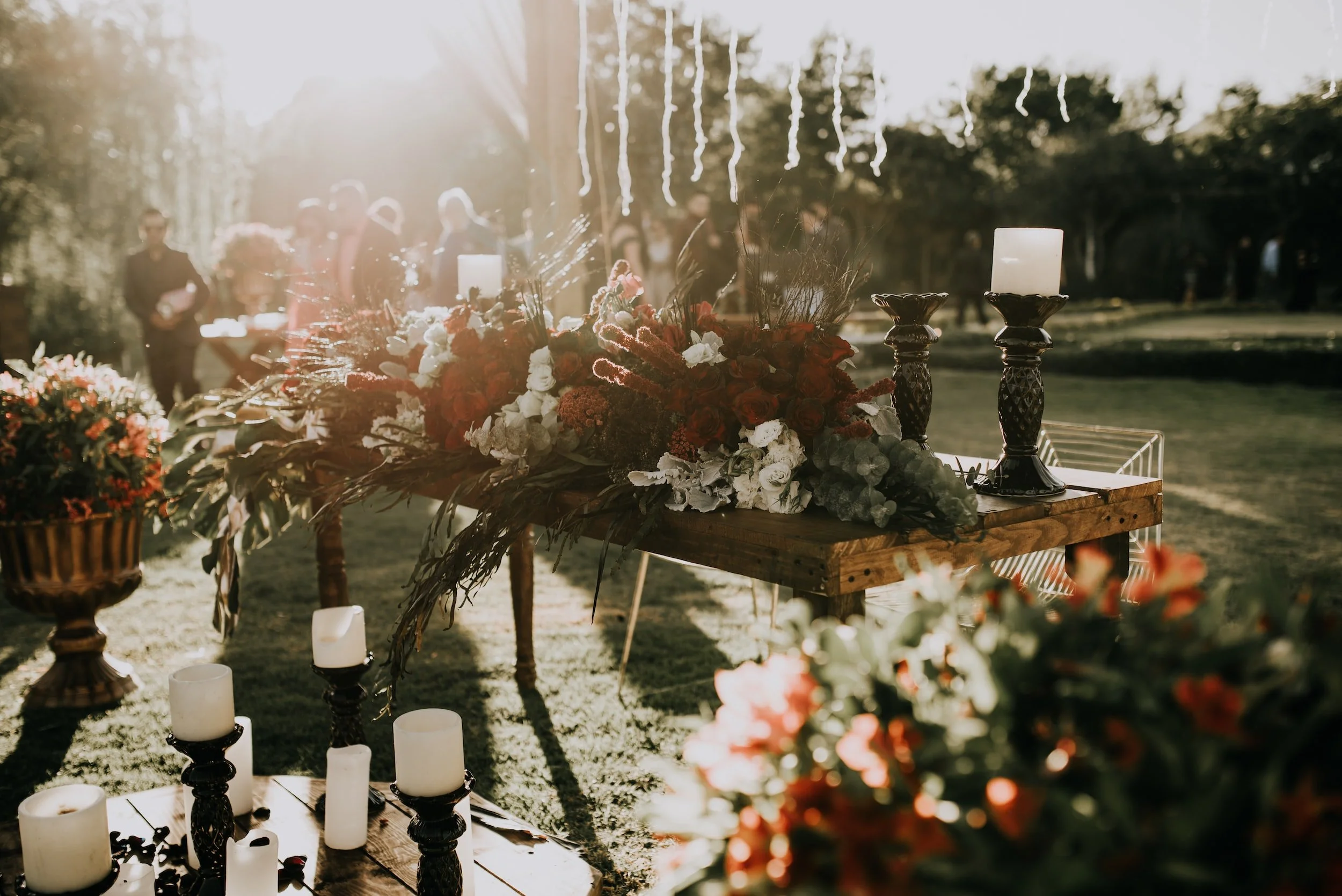 Decorative floral table arrangement with candles at an outdoor event, sunlight shining through trees in the background.