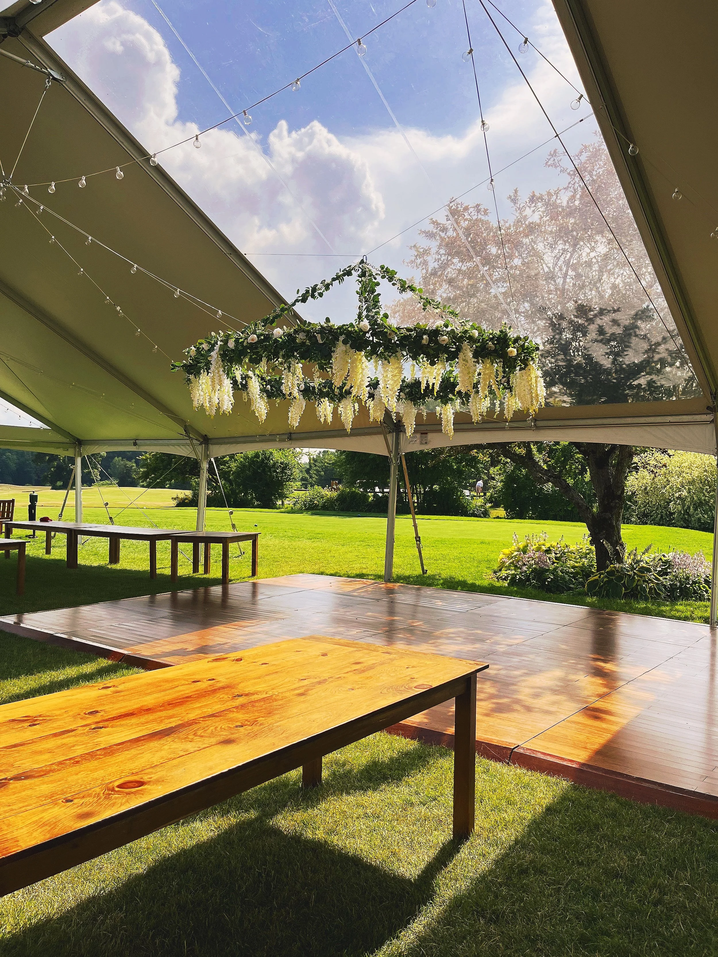 Outdoor event tent with string lights and a floral hanging decoration, set on a grassy field with trees and a cloudy sky in the background.
