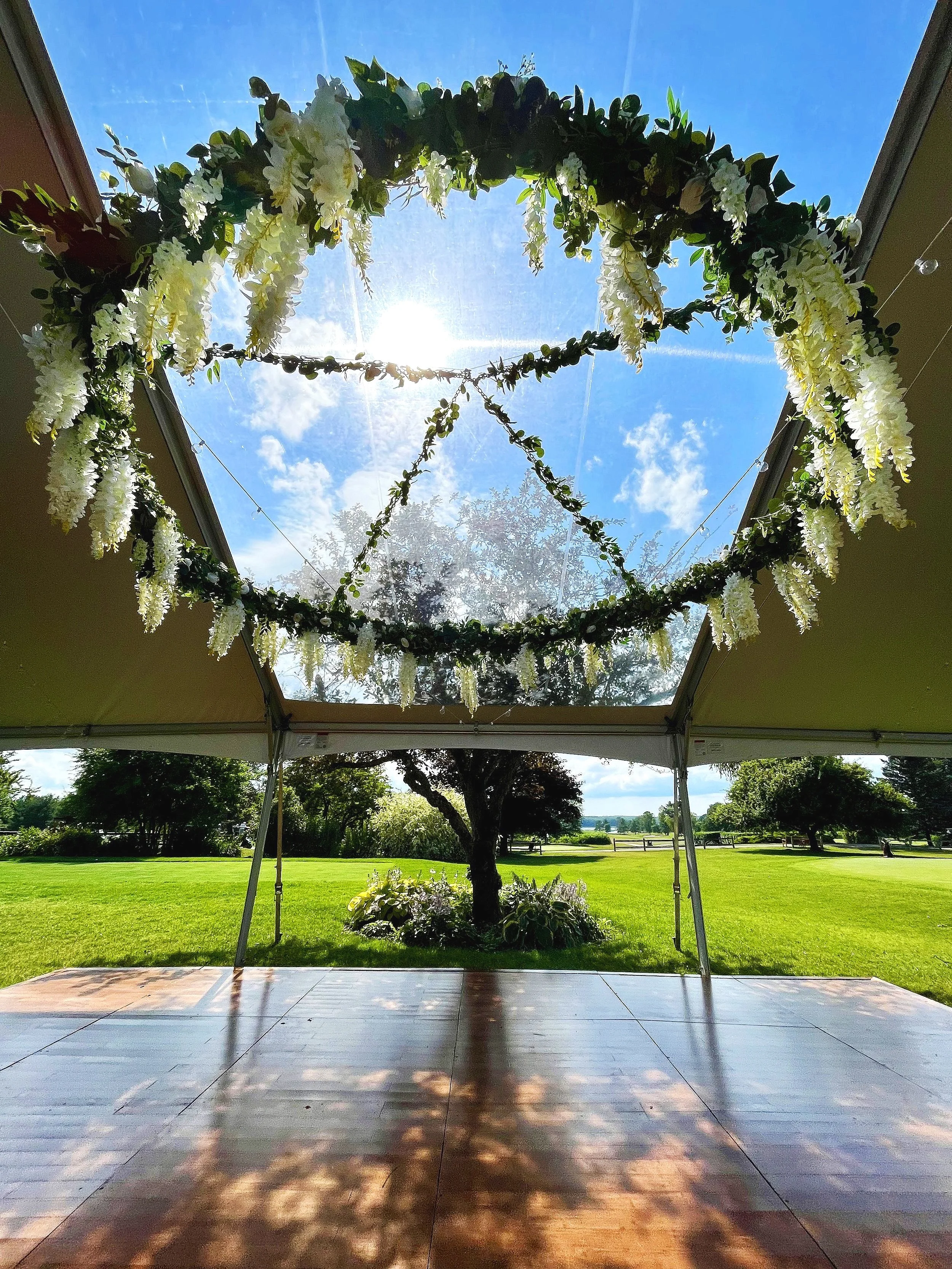 View of a decorated outdoor canopy with hanging floral wreaths, overlooking a grassy park with trees and a bright blue sky.