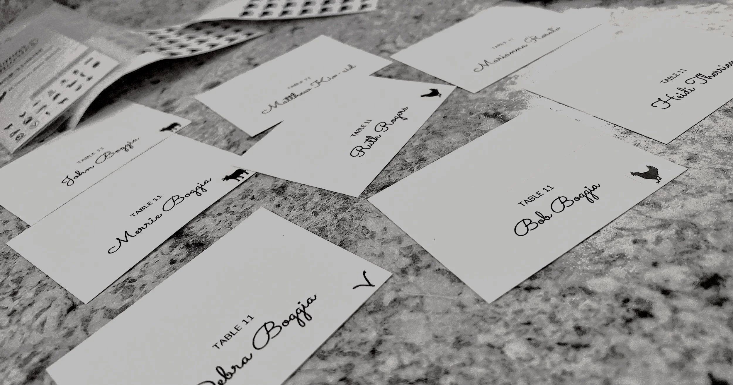 Wedding place cards with the names Mona Boegia and Rhian Boegia, and black dog illustrations, arranged on a table with stacks of paper and a calculator nearby.