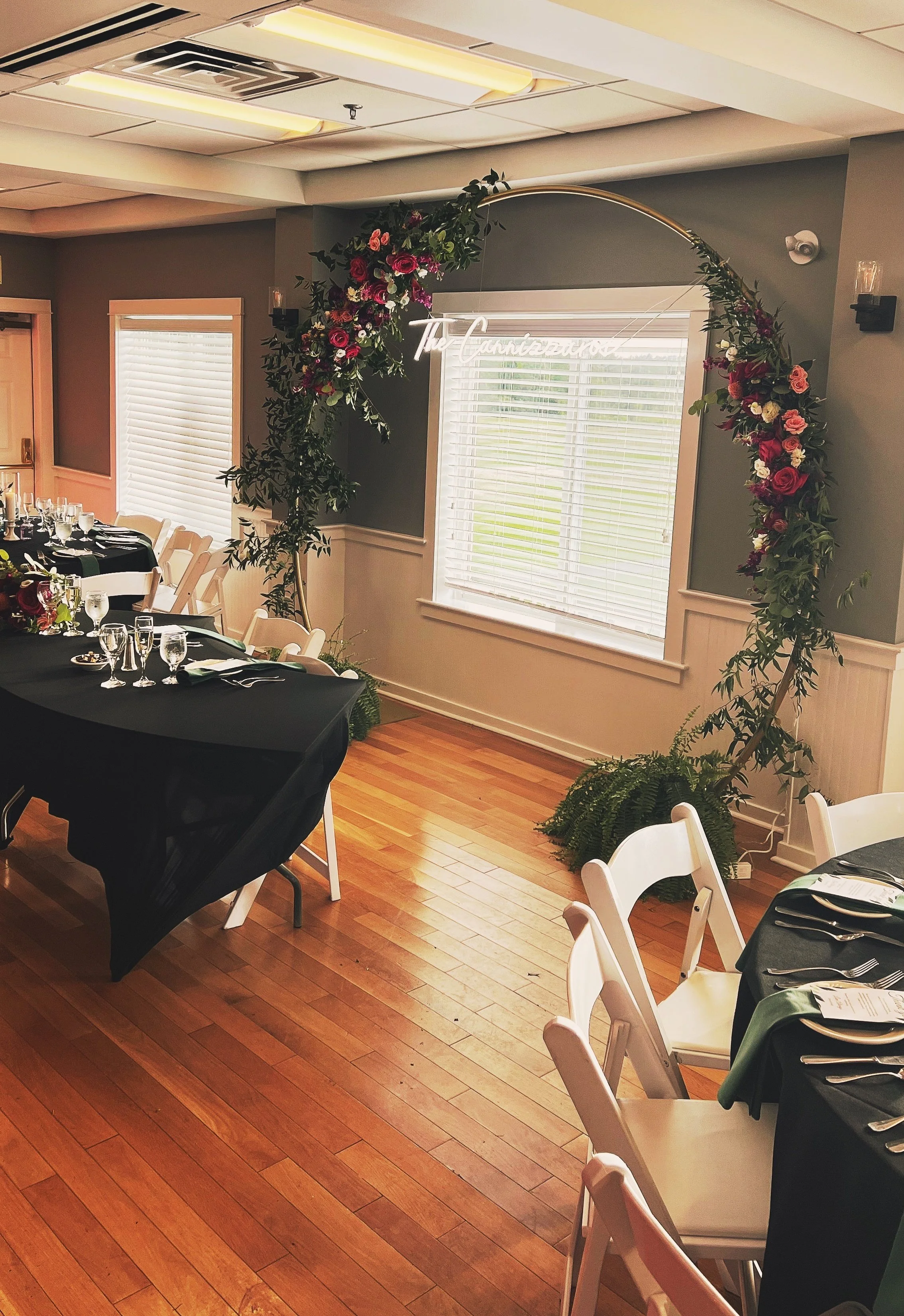 Decorated banquet hall with floral arch, circular tables with black tablecloths, white chairs, set with glassware, silverware, and menus, with natural light from windows.