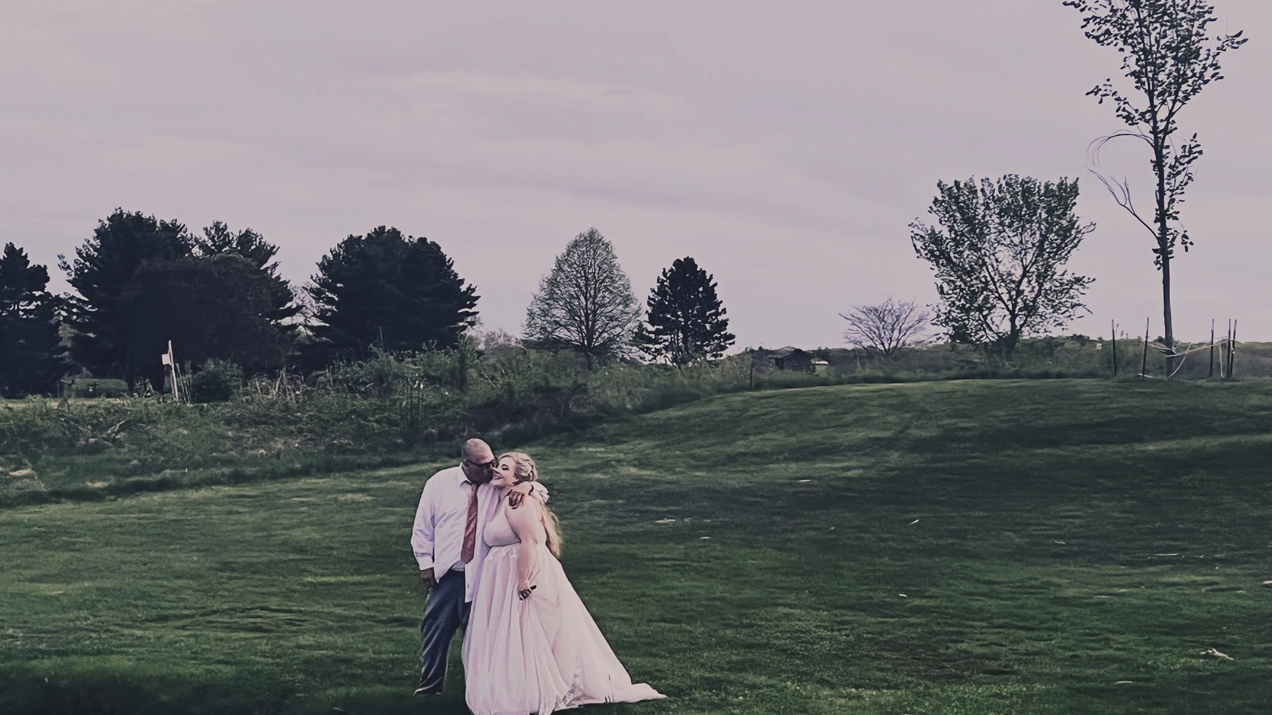 A couple, the man in a white shirt and tie, and the woman in a long, flowing dress, sharing a kiss on a grassy field with trees and overcast sky in the background.