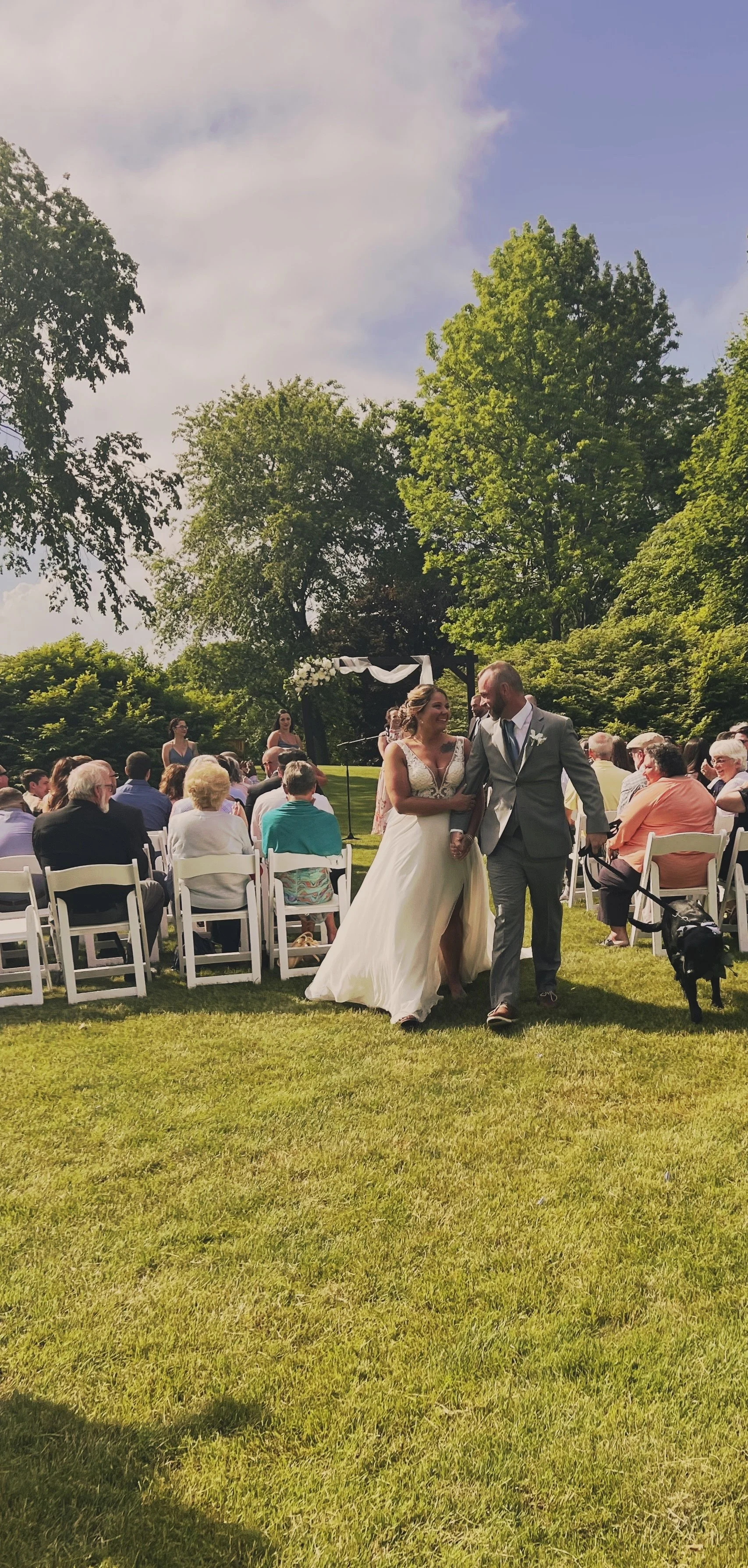 A bride and groom walking together at their outdoor wedding, surrounded by seated guests and lush green trees under a partly cloudy sky.