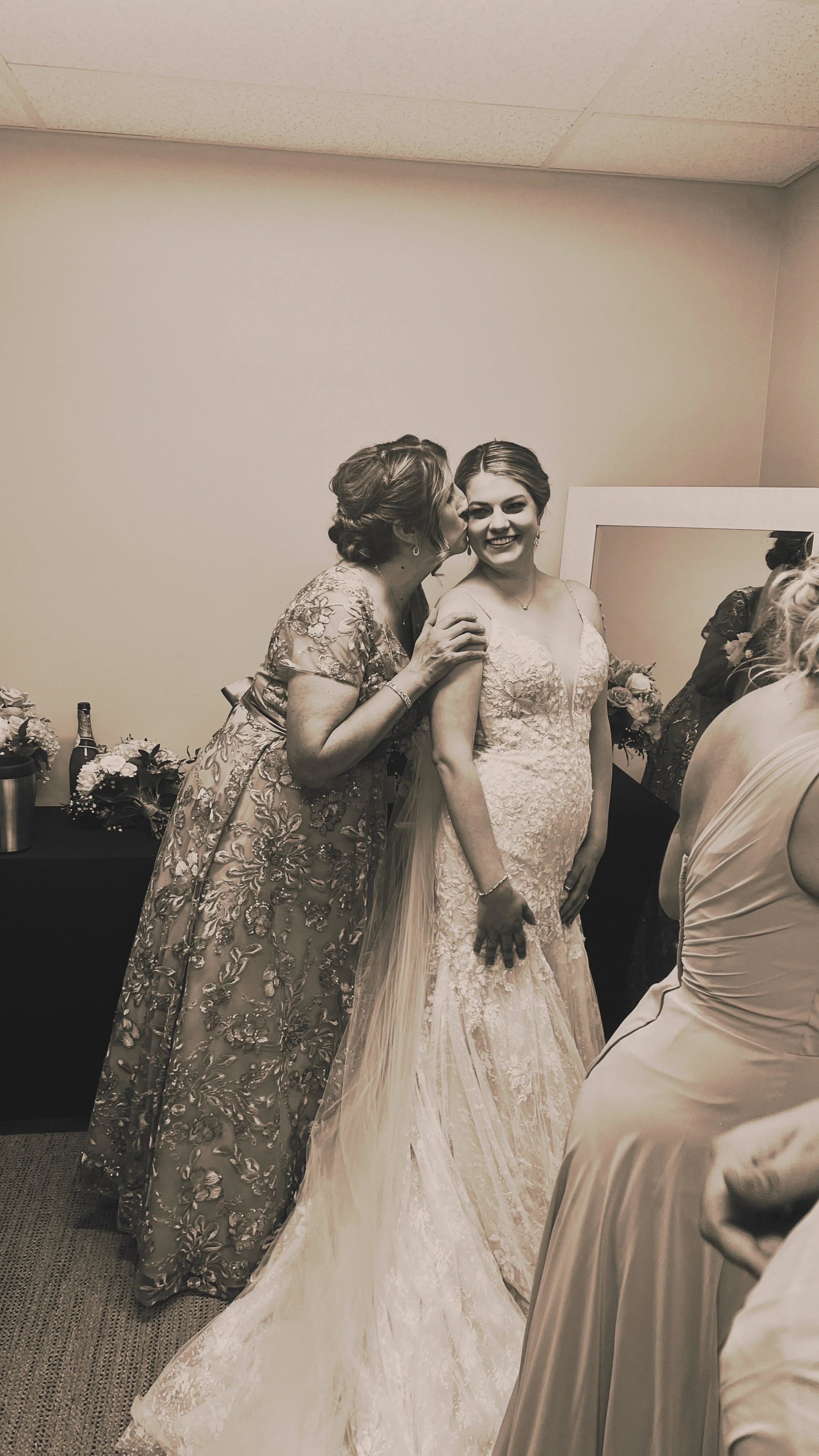 A bride in a lace wedding gown being kissed on the cheek by an older woman in a floral dress, standing in a dressing room with friends and flowers in the background.