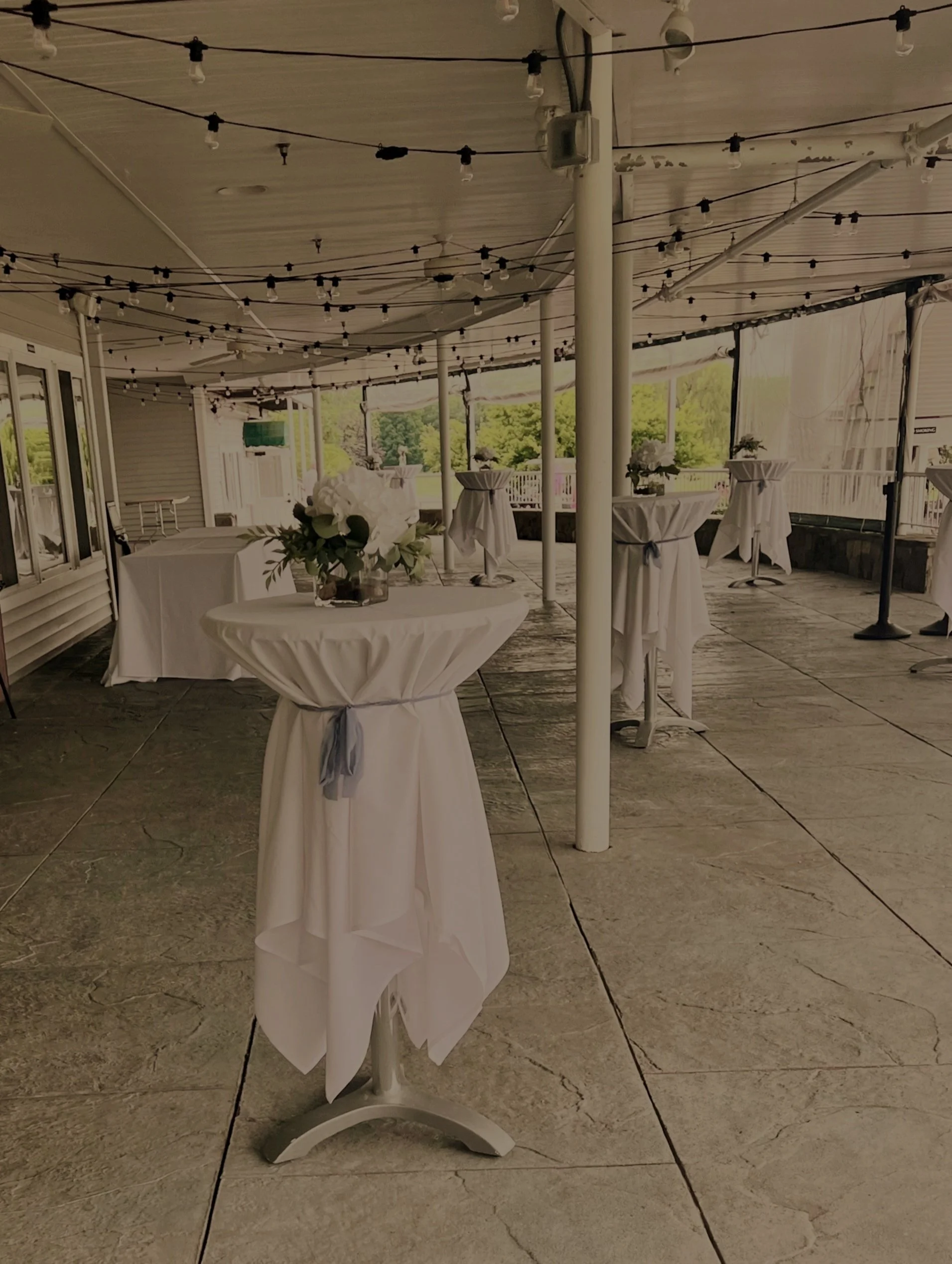 Empty outdoor event space with high-top tables decorated with white tablecloths and floral arrangements, hanging string lights, and a view of trees in the background.