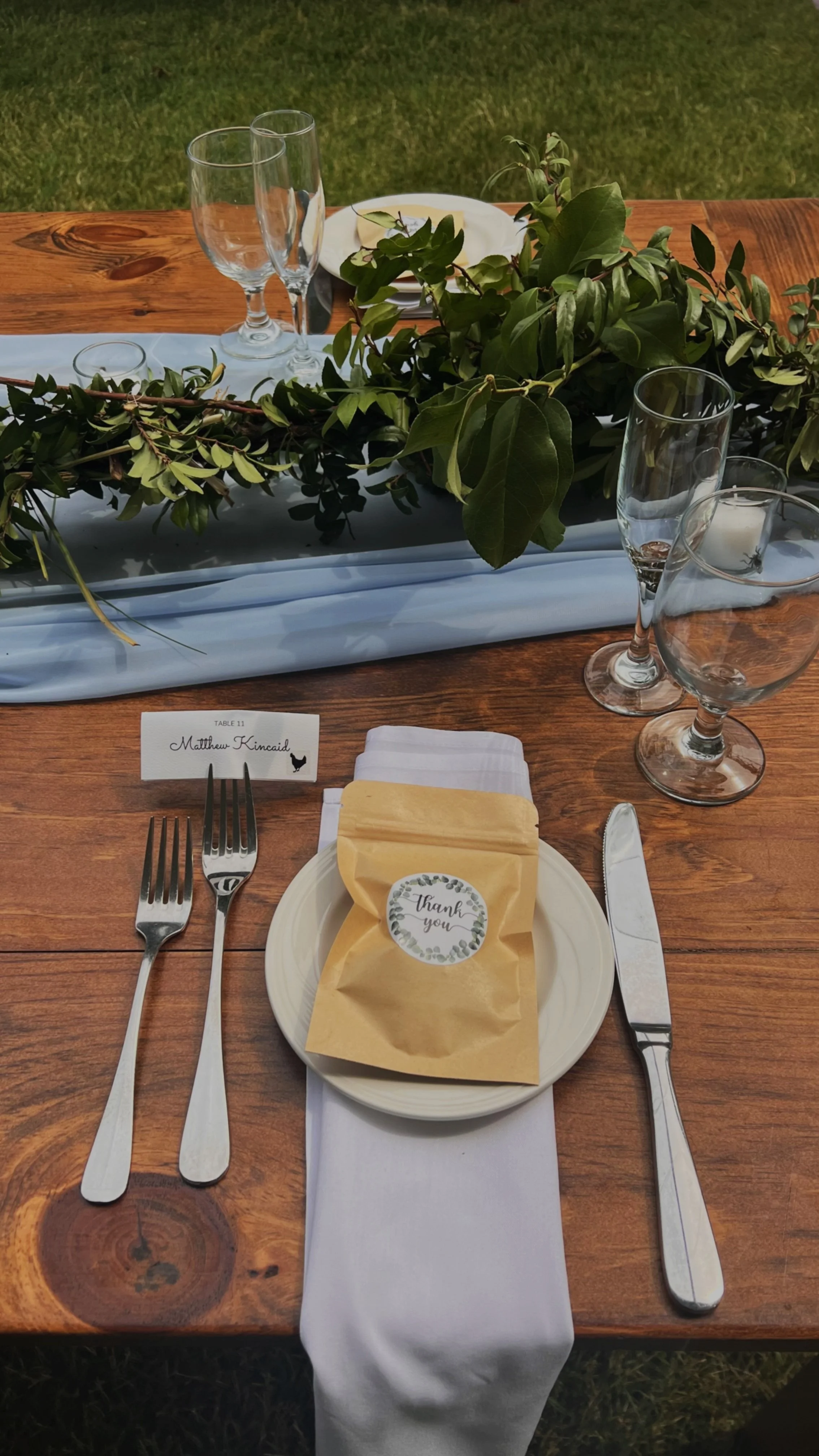 A formal outdoor table setting with a wooden table, white napkin, name tag reading 'Matthew Kincaid', glassware including two wine glasses, a plate with a yellow bag labeled 'Thank You', a knife, a fork, a spoon, and a decorative green foliage center