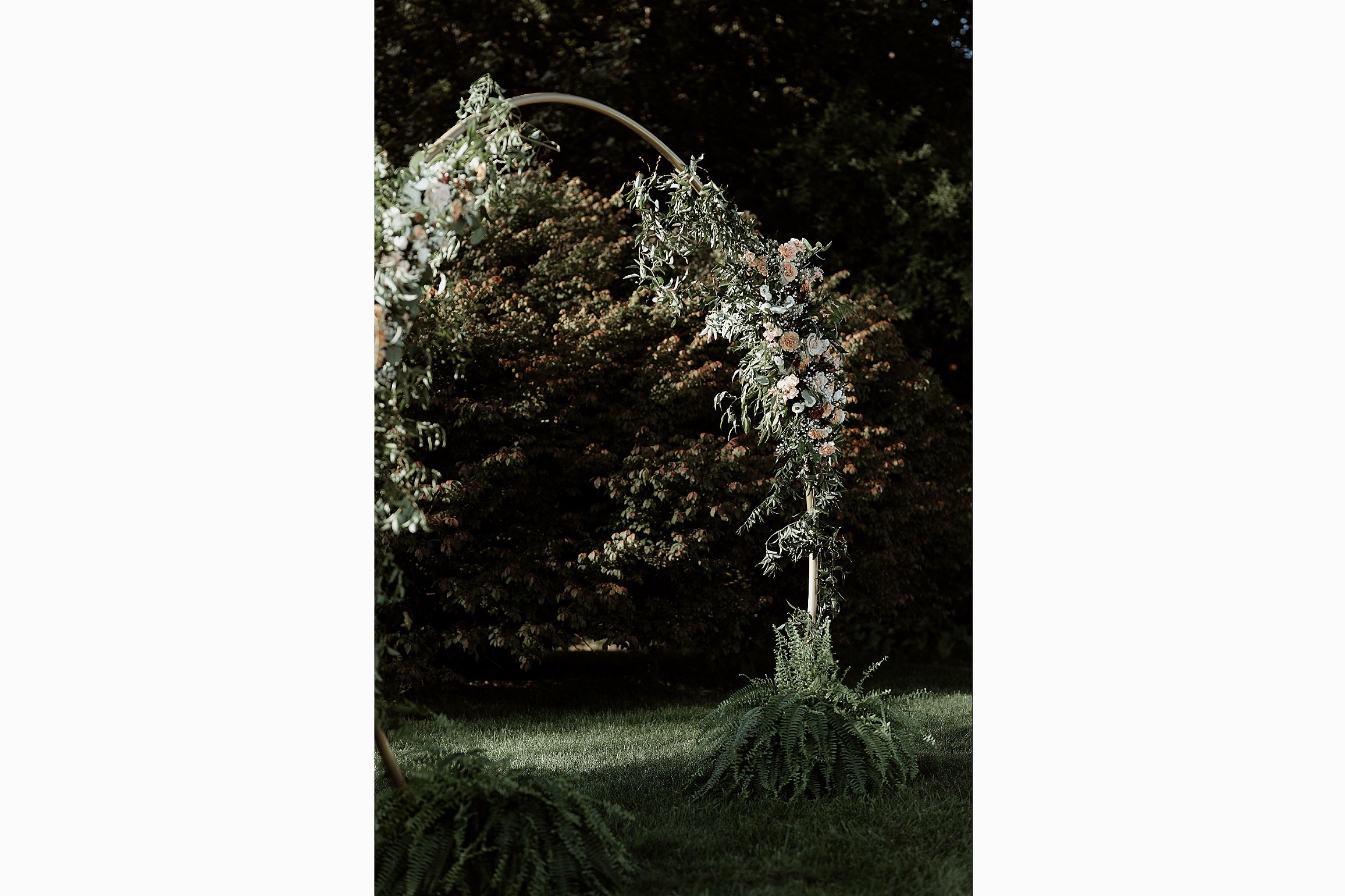 Floral wedding arch decorated with white, pink, and peach flowers and greenery, standing outdoors on a grassy area with trees in the background.