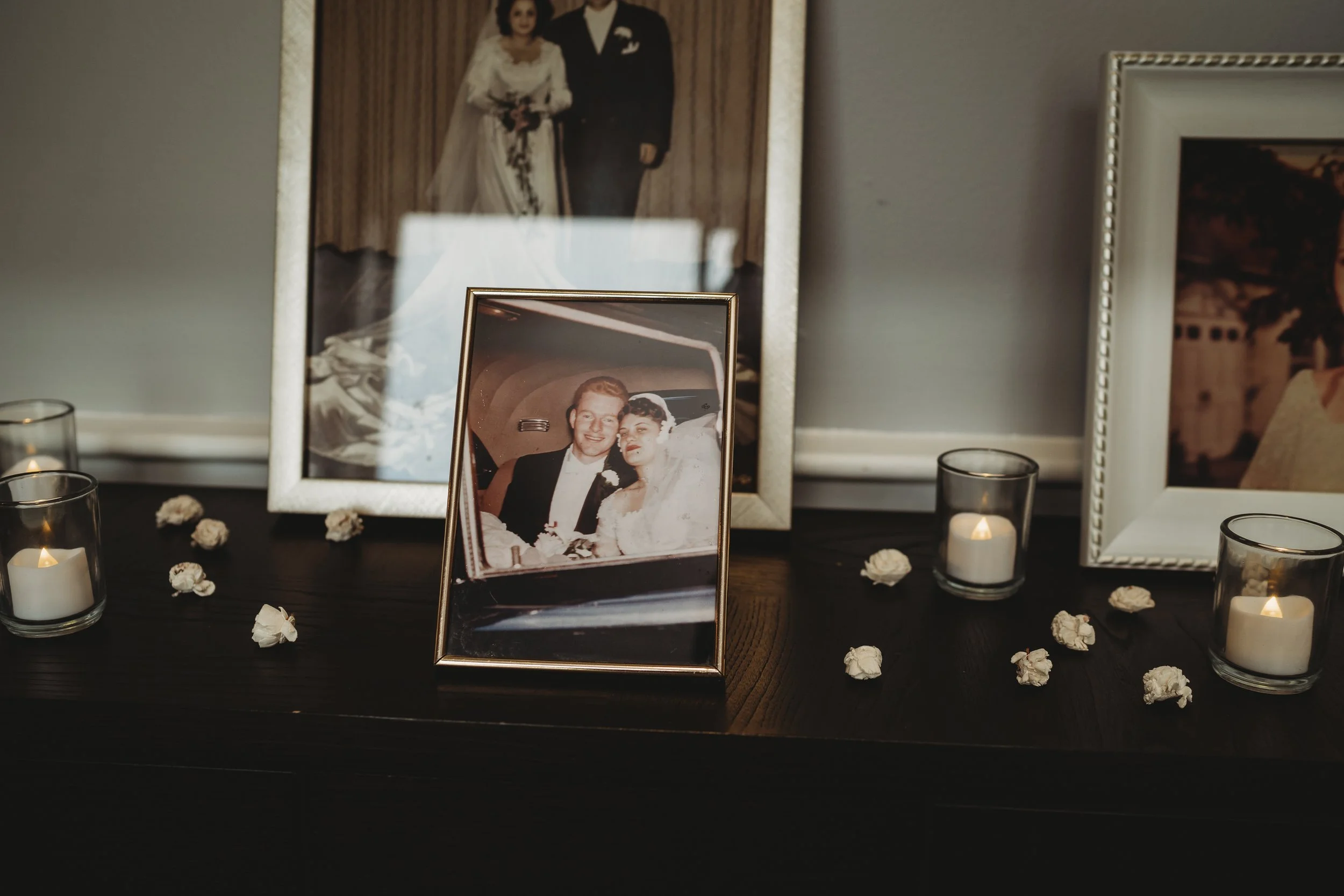 Photographs of a wedding displayed on a dark wooden surface, including a framed picture of a bride and groom, with candles around and scattered white flower petals.