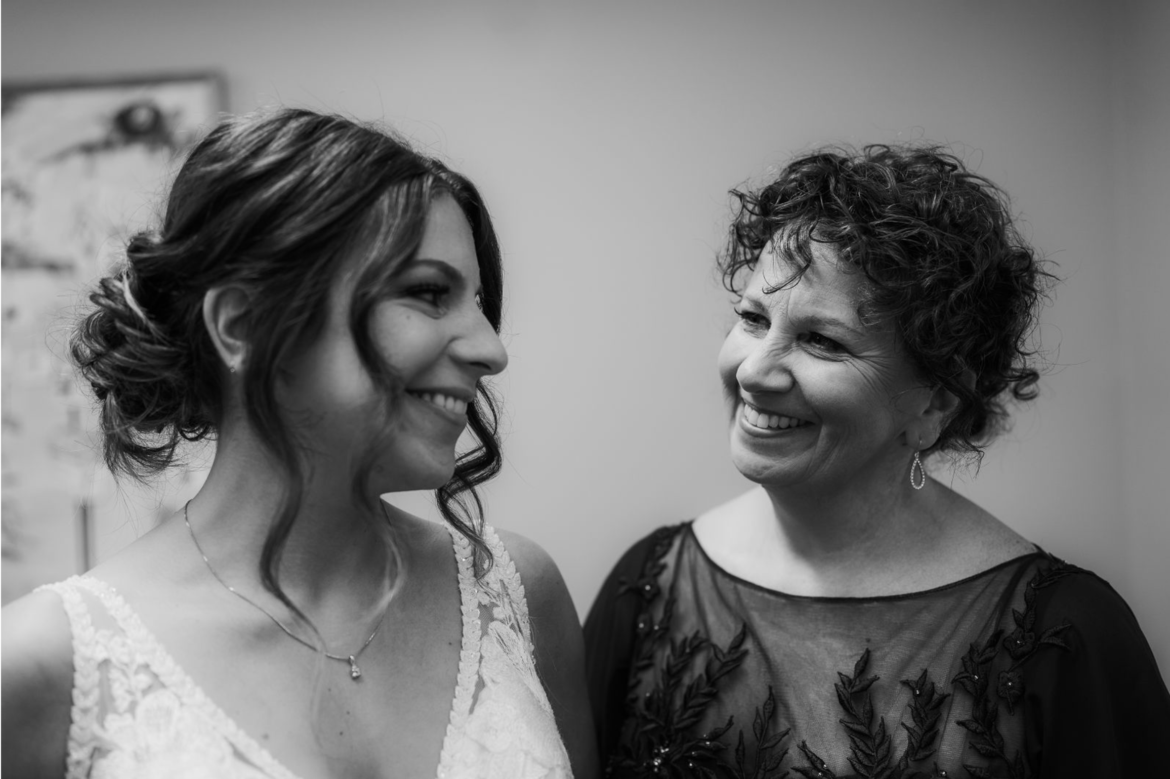 A young woman in a wedding dress smiling at an older woman in a black dress, both face to face in an intimate moment.