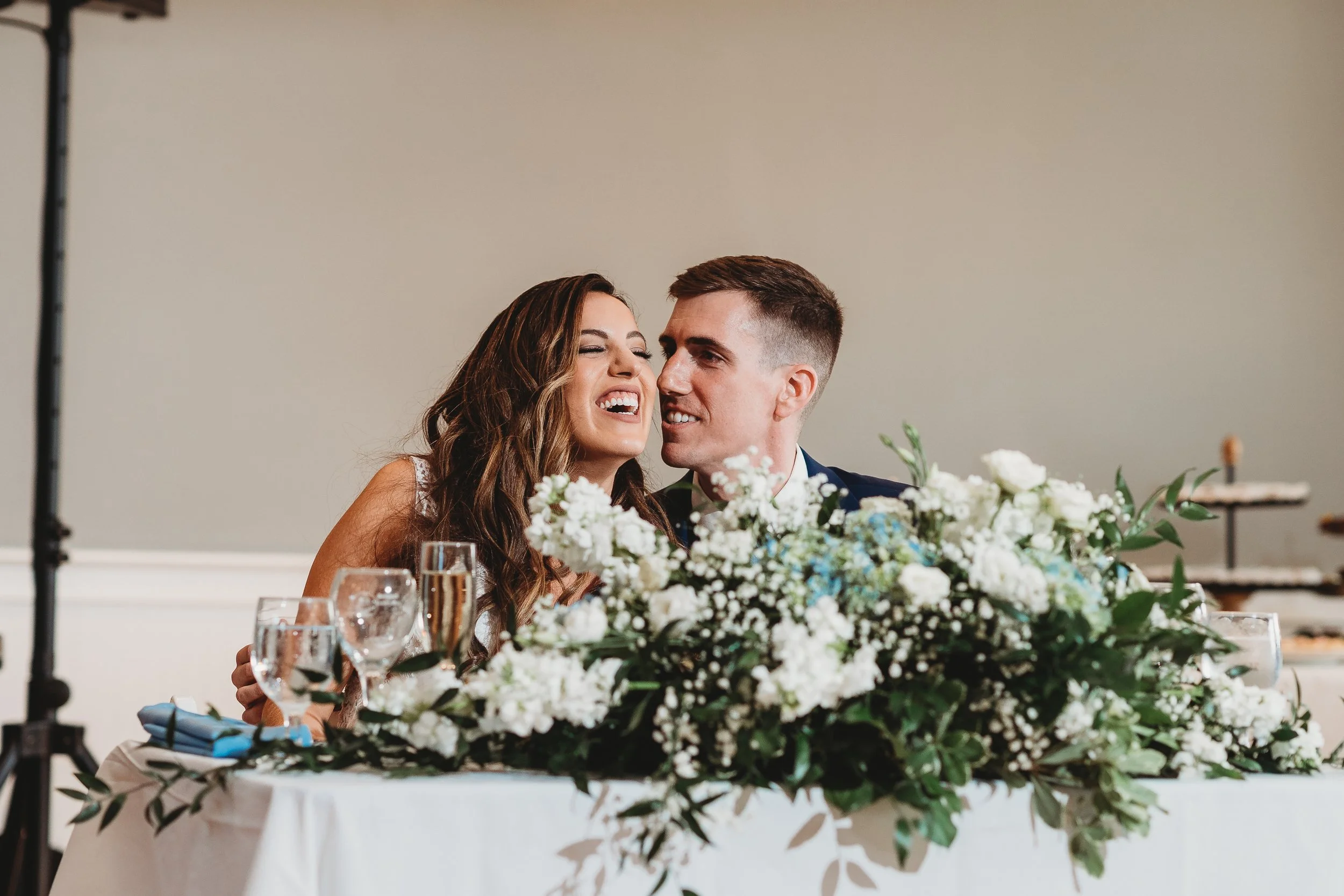 A bride and groom sitting at a wedding reception table decorated with a large floral arrangement of white flowers and greenery, smiling and leaning close to each other.