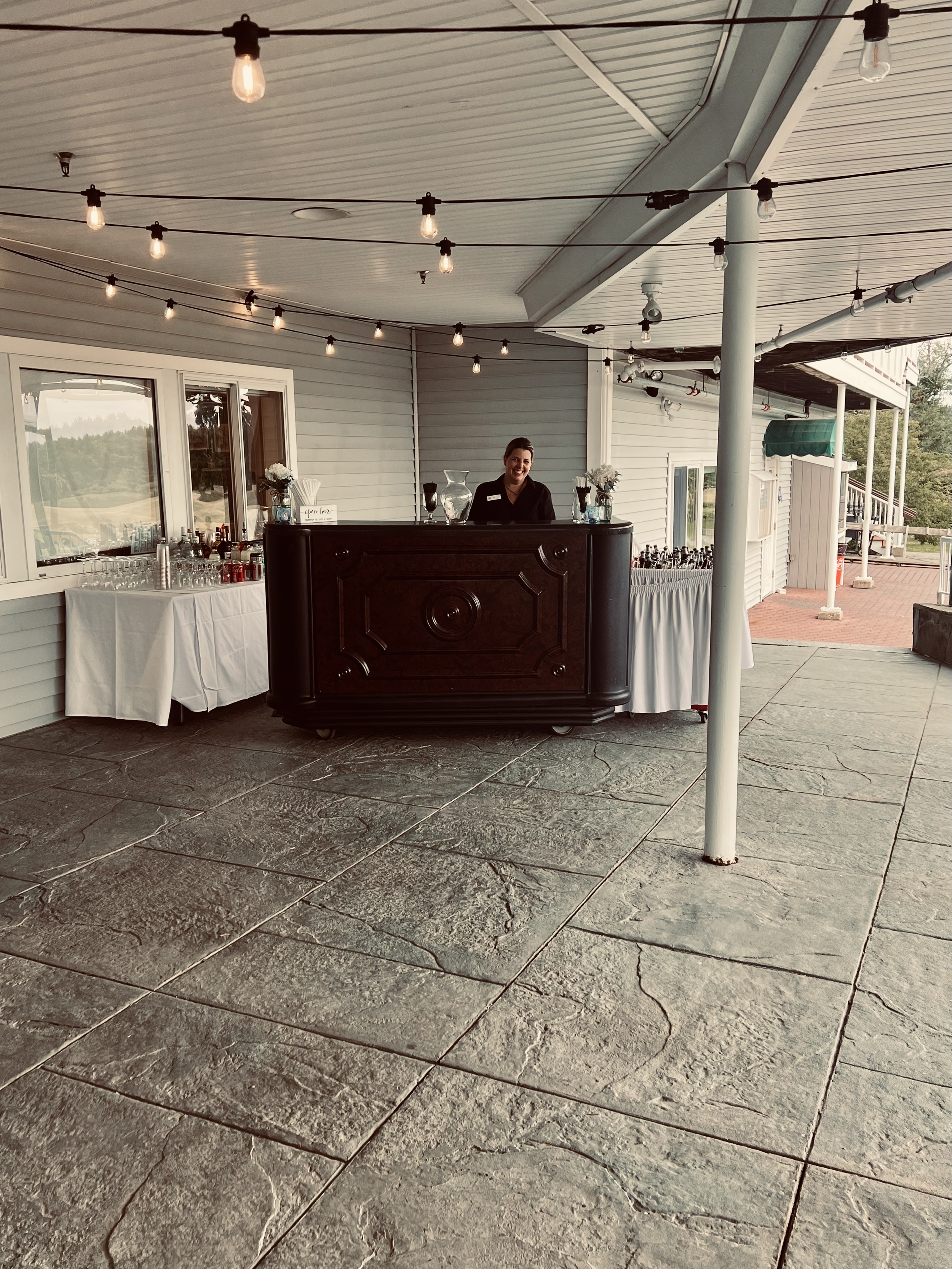 A bartender standing behind a bar at an outdoor event space with string lights overhead and tables with drinks on either side.
