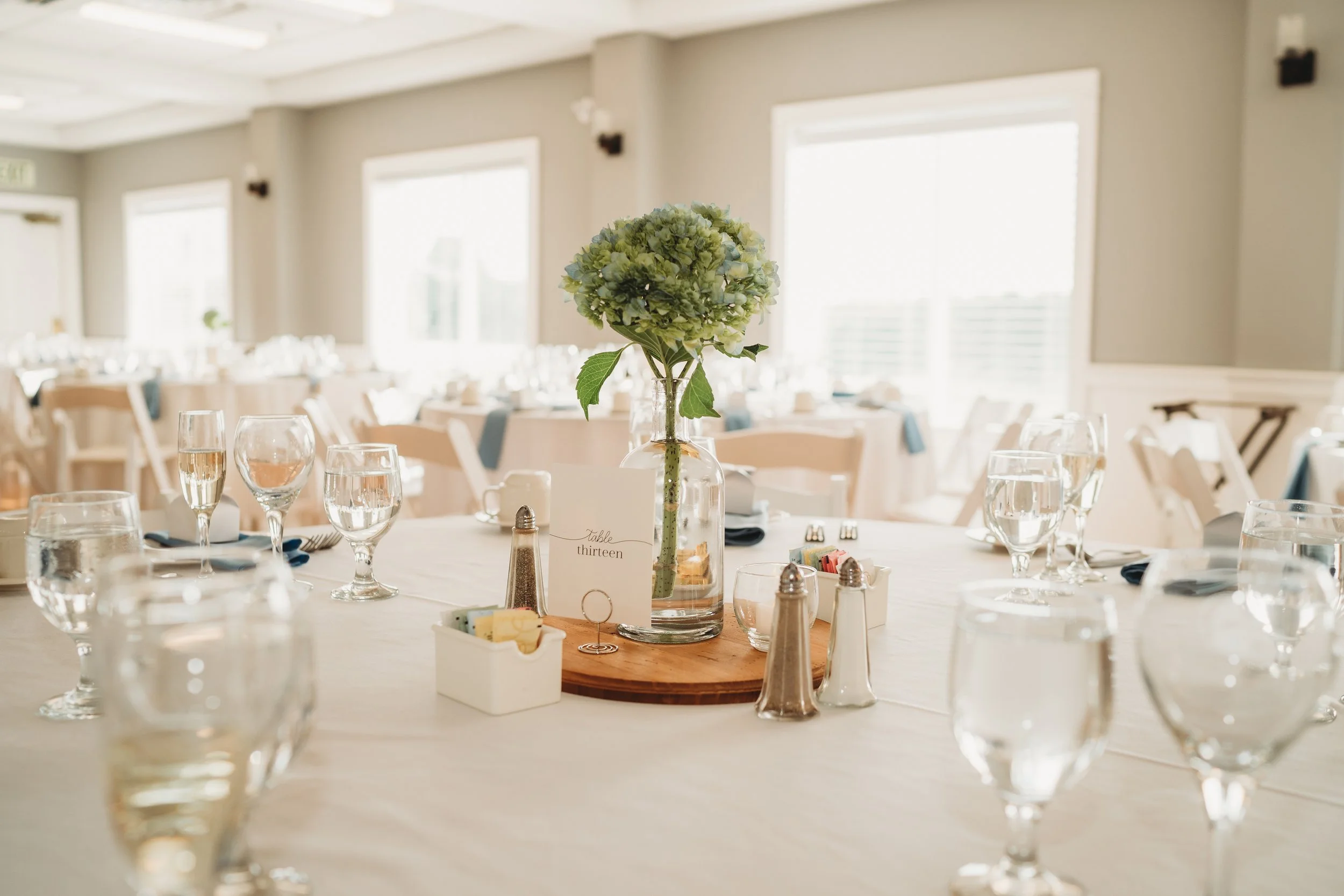 Wedding reception table decorated with a green hydrangea centerpiece, wine glasses, water glasses, salt and pepper shakers, butter packets, and a table number card, in a bright room with large windows.