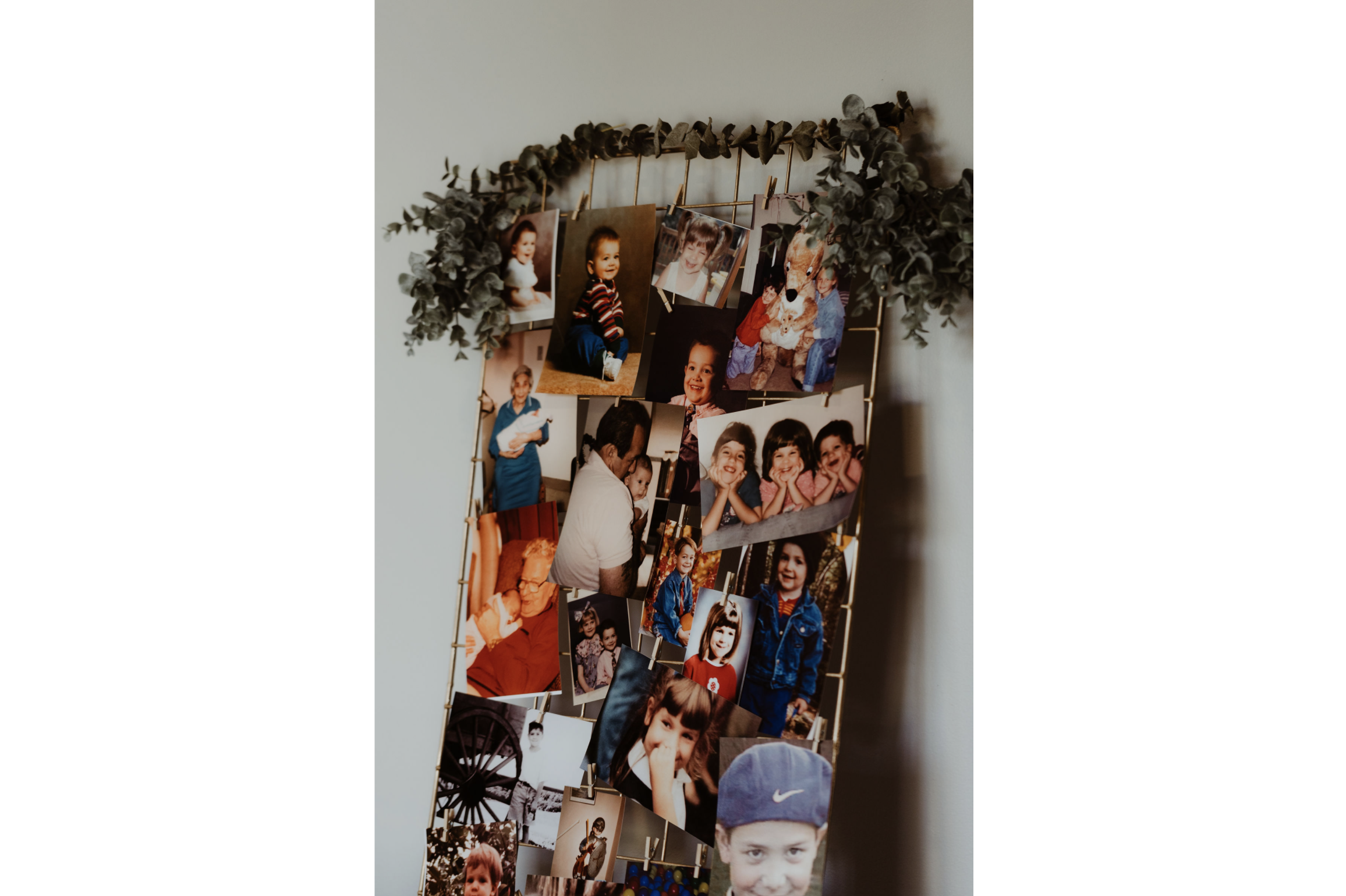 A photo collage of various childhood photographs hanging on a gold wire grid decorated with greenery on a light-colored wall.