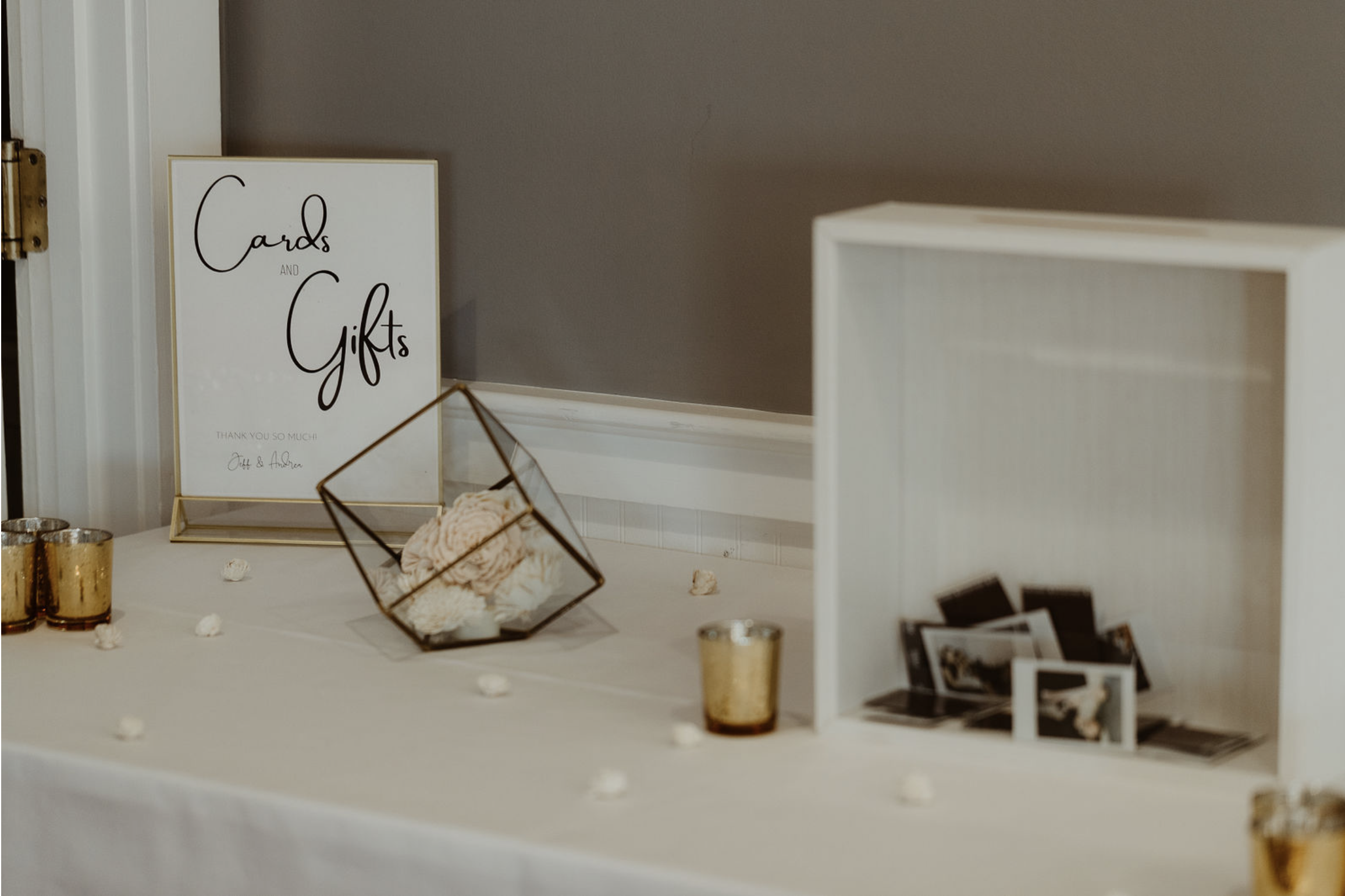 Decorative table with a sign reading 'Cards and Gifts', a glass terrarium containing flowers, small candles, and a box with photographs at an event.