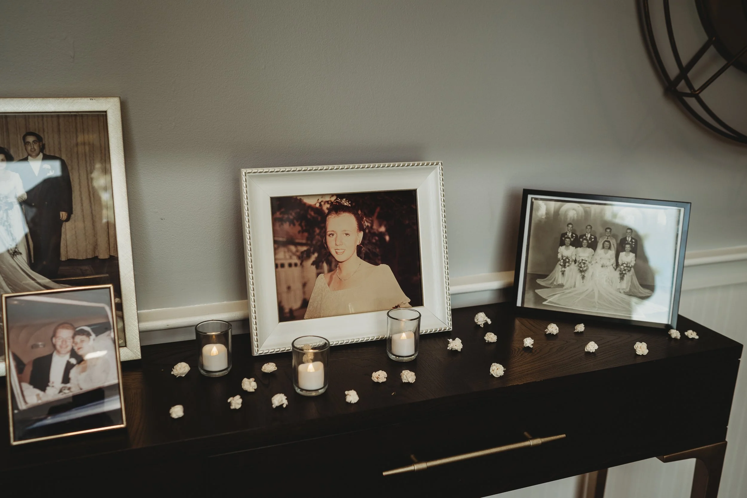 A collection of framed family photographs and small white flowers on a dark wooden table, with three lit candles in glass holders and a gray wall in the background.