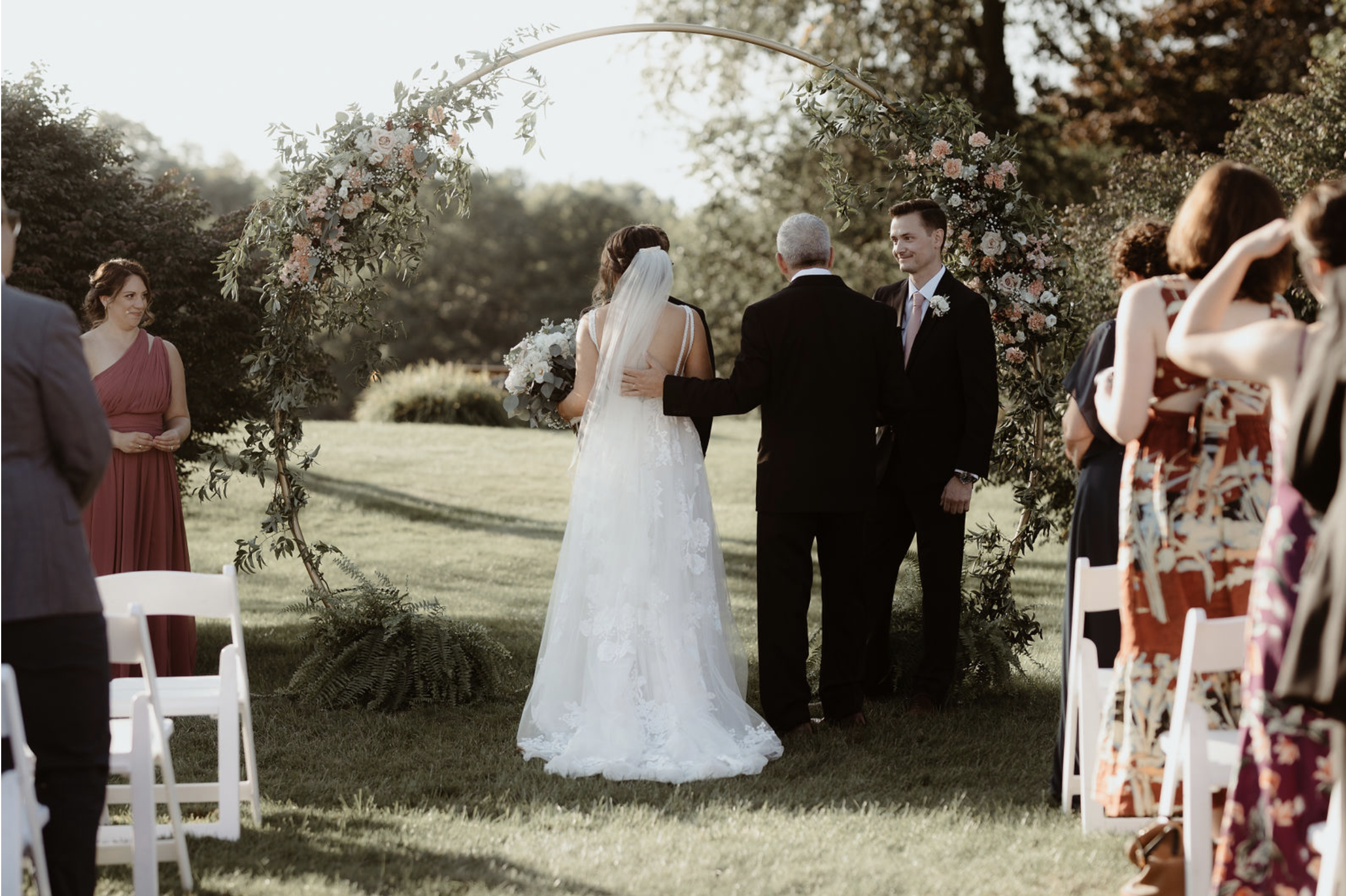 Bride and groom exchange vows under an outdoor floral arch as guests look on.