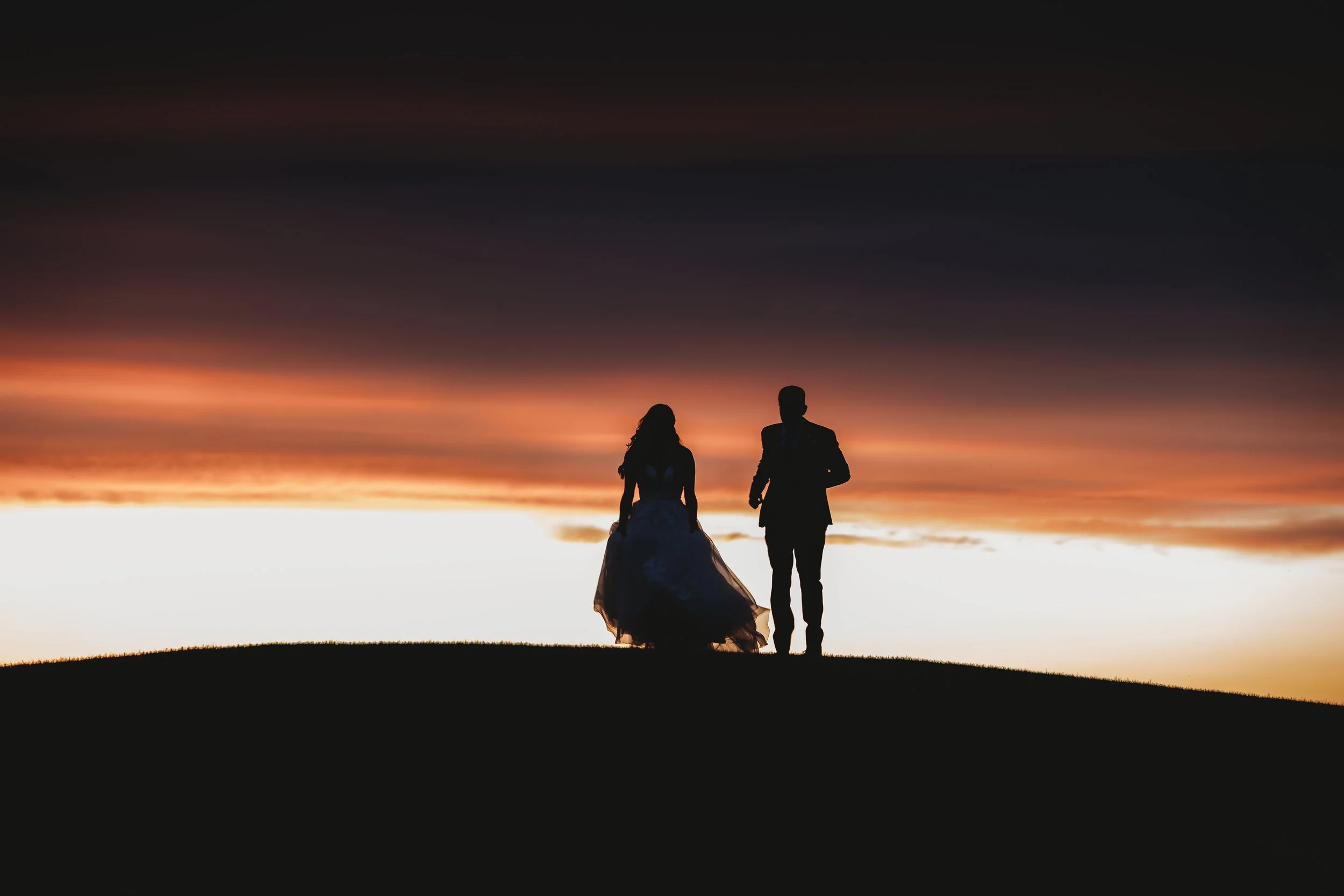 Silhouettes of a man and woman walking together on a hill at sunset with dramatic orange and purple sky.