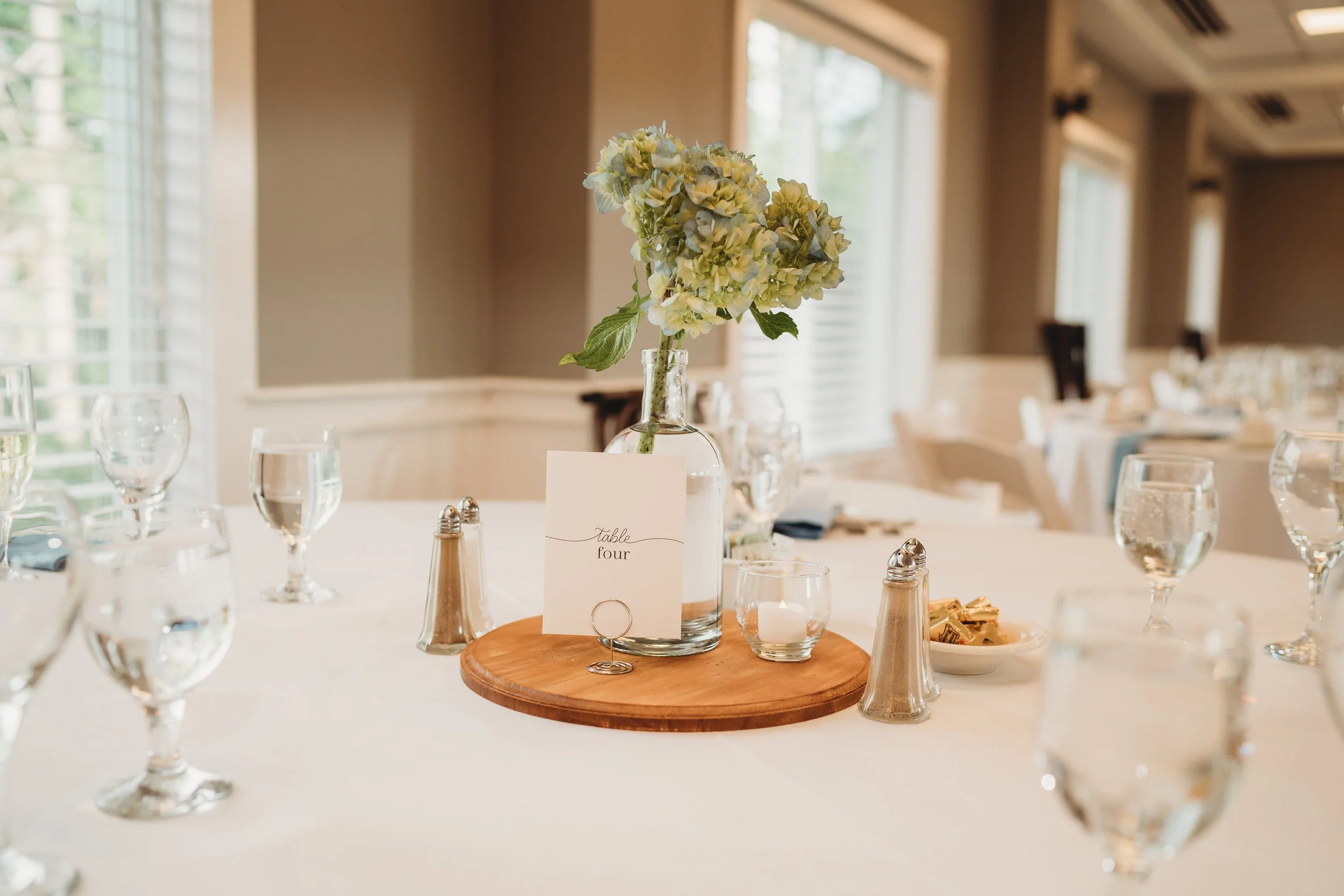 A wedding reception table with a white tablecloth, a centerpiece of green and white hydrangeas in a glass vase, a candle, salt and pepper shakers, a card indicating 'table four,' and a small bowl of candy. Multiple glasses of water are arranged aroun