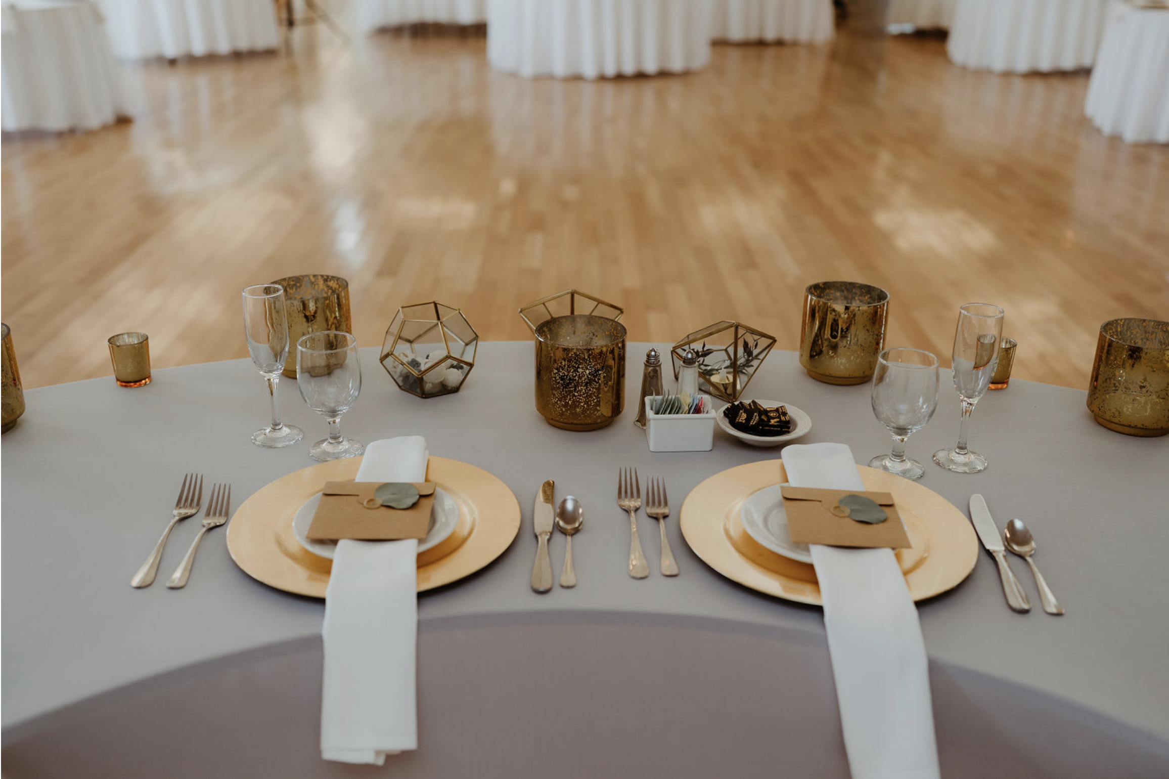 Elegant banquet table set with gold chargers, white plates, gold flatware, white napkins, and place cards. Centerpieces include gold candle holders, geometric glass terrariums, and a small container with sugar packets. The background shows a wooden d