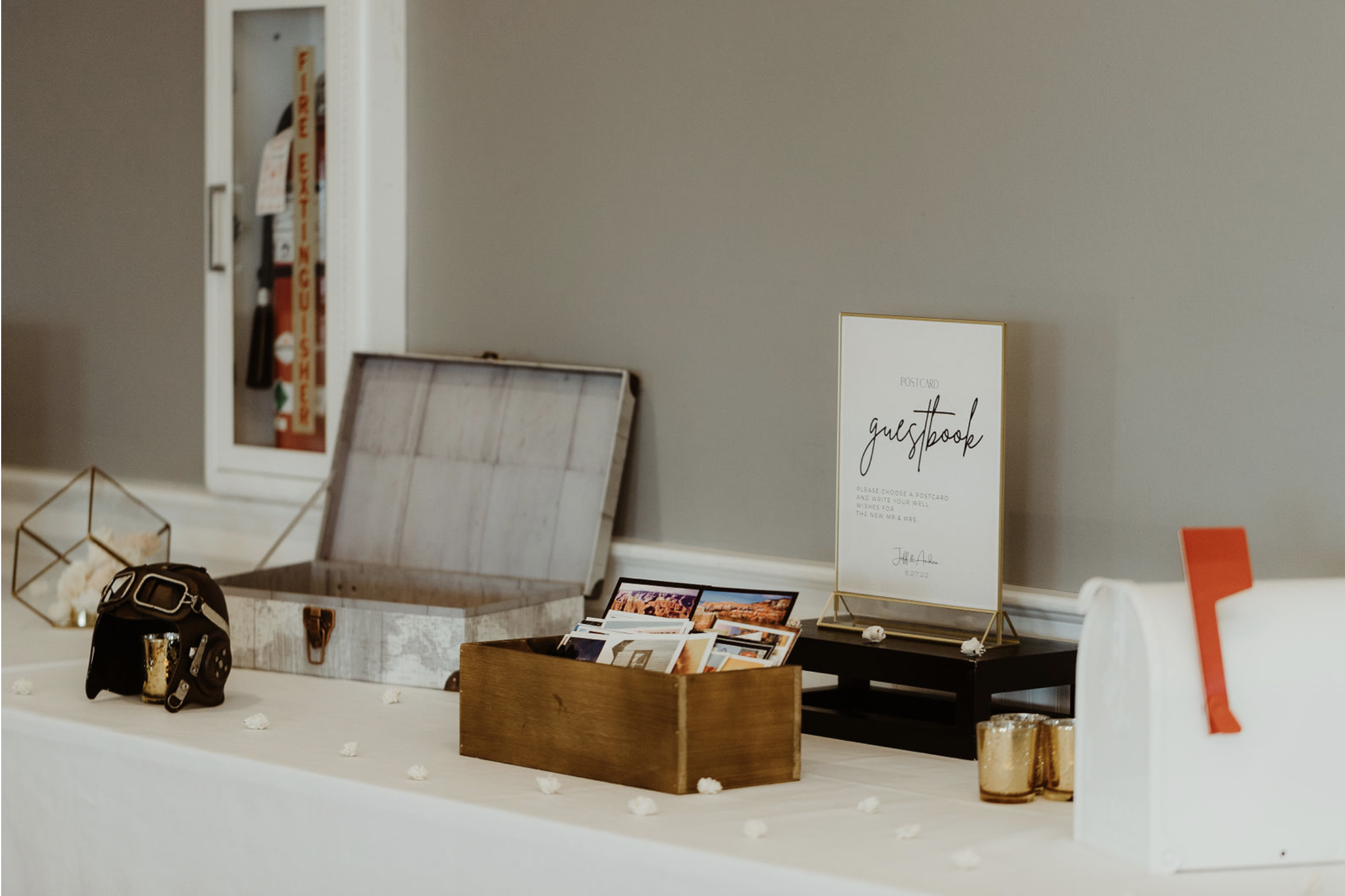Table with guestbook sign, postcards, a wooden box with photos, decorative candle holders, a vintage style camera, and a geometric photo display, set up for a wedding or event.