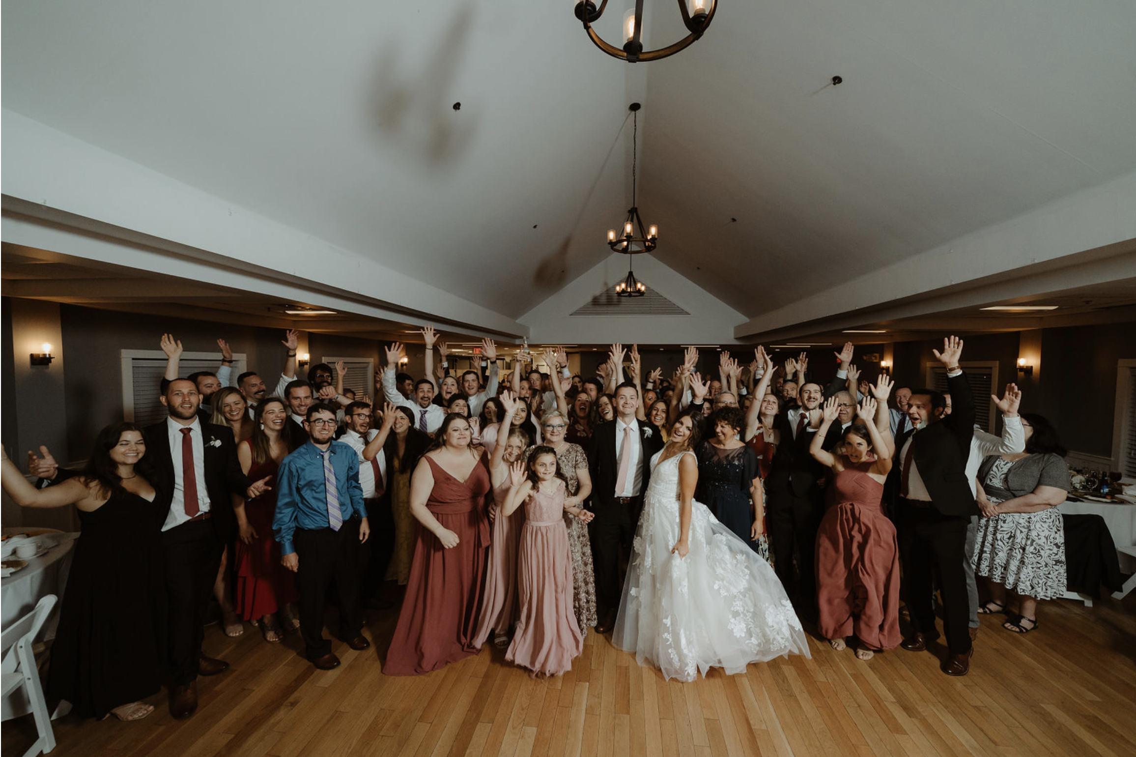 Group of people, including a bride and groom, celebrating at a wedding reception with many guests raising their hands in the air inside a banquet hall.