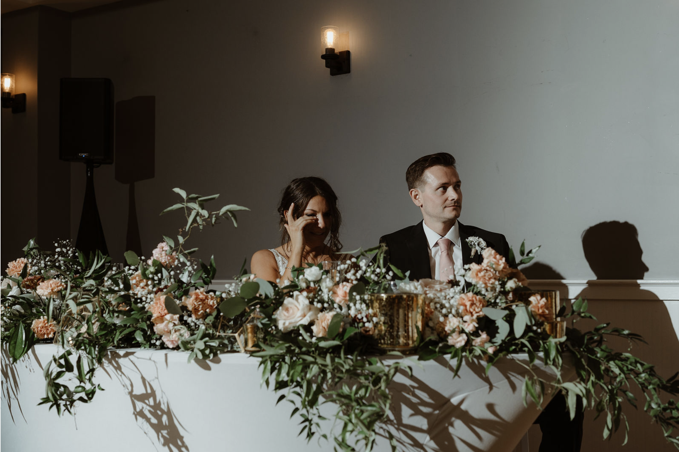 A wedding reception table decorated with pink and white flowers and greenery, with a bride and groom sitting behind it. The bride is wiping tears from her eyes, and the groom is looking to the side.