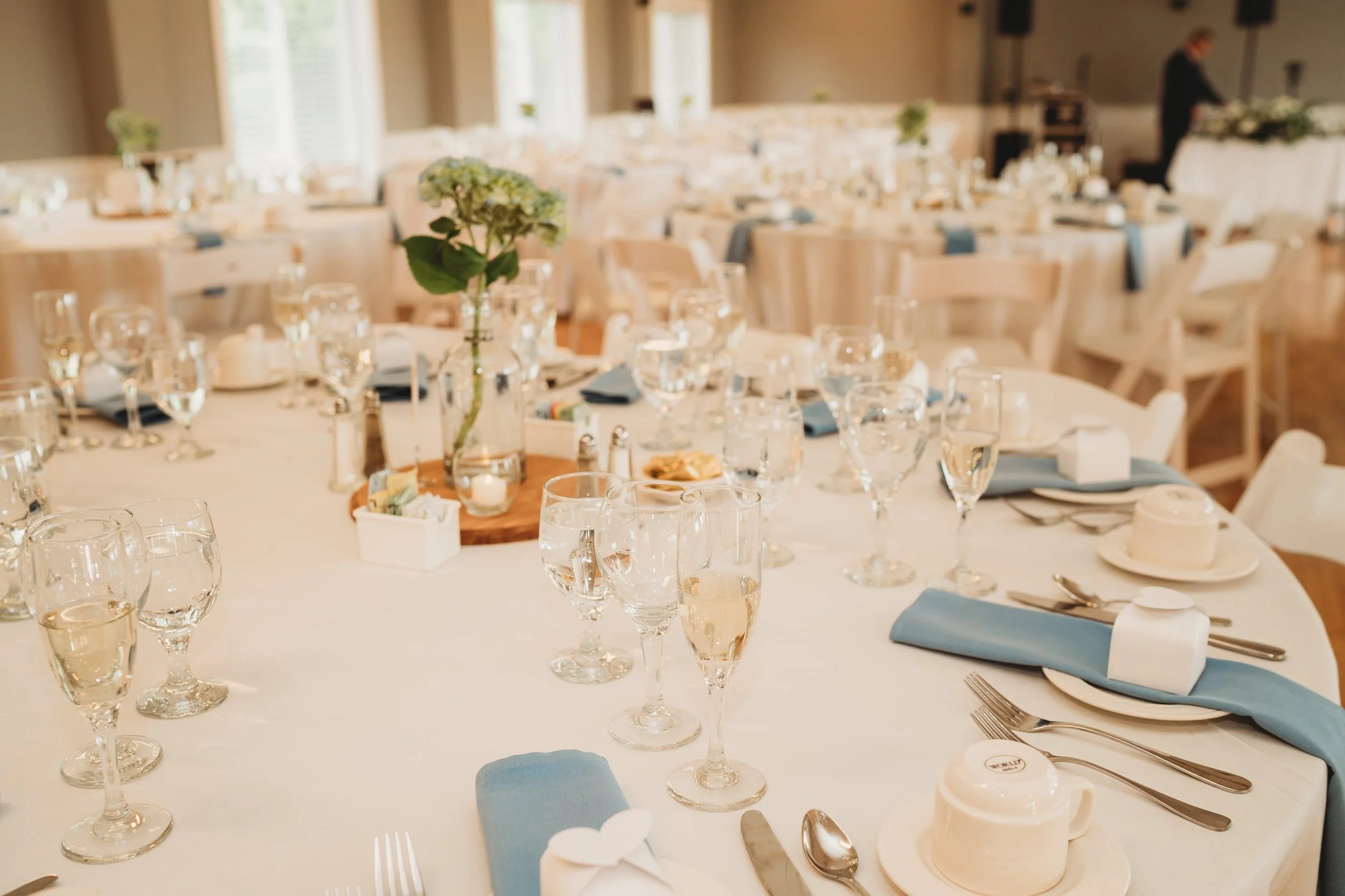 Elegant banquet table set with wine glasses, water glasses, plates, silverware, blue napkins, and a centerpiece with a hydrangea flower in a glass vase, in a well-lit room.