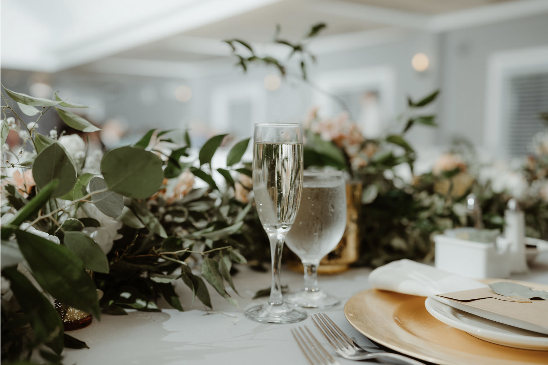 A table setting with a glass of champagne, a glass of water, a plate with napkin, and a gold charger, decorated with greenery and floral arrangements in the background.