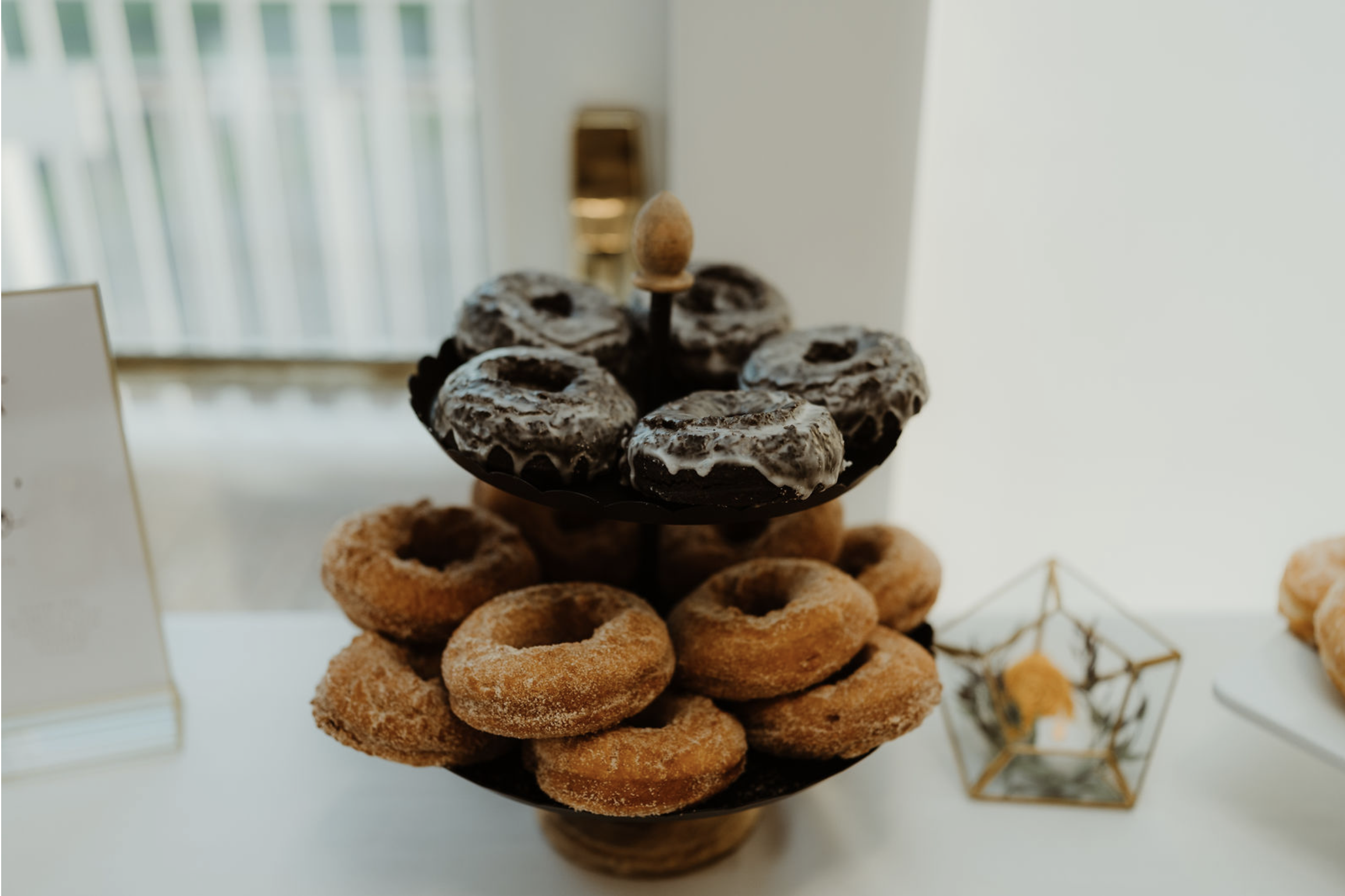A two-tiered stand holding glazed donuts and cinnamon sugar donuts, situated on a white table in a brightly lit room.