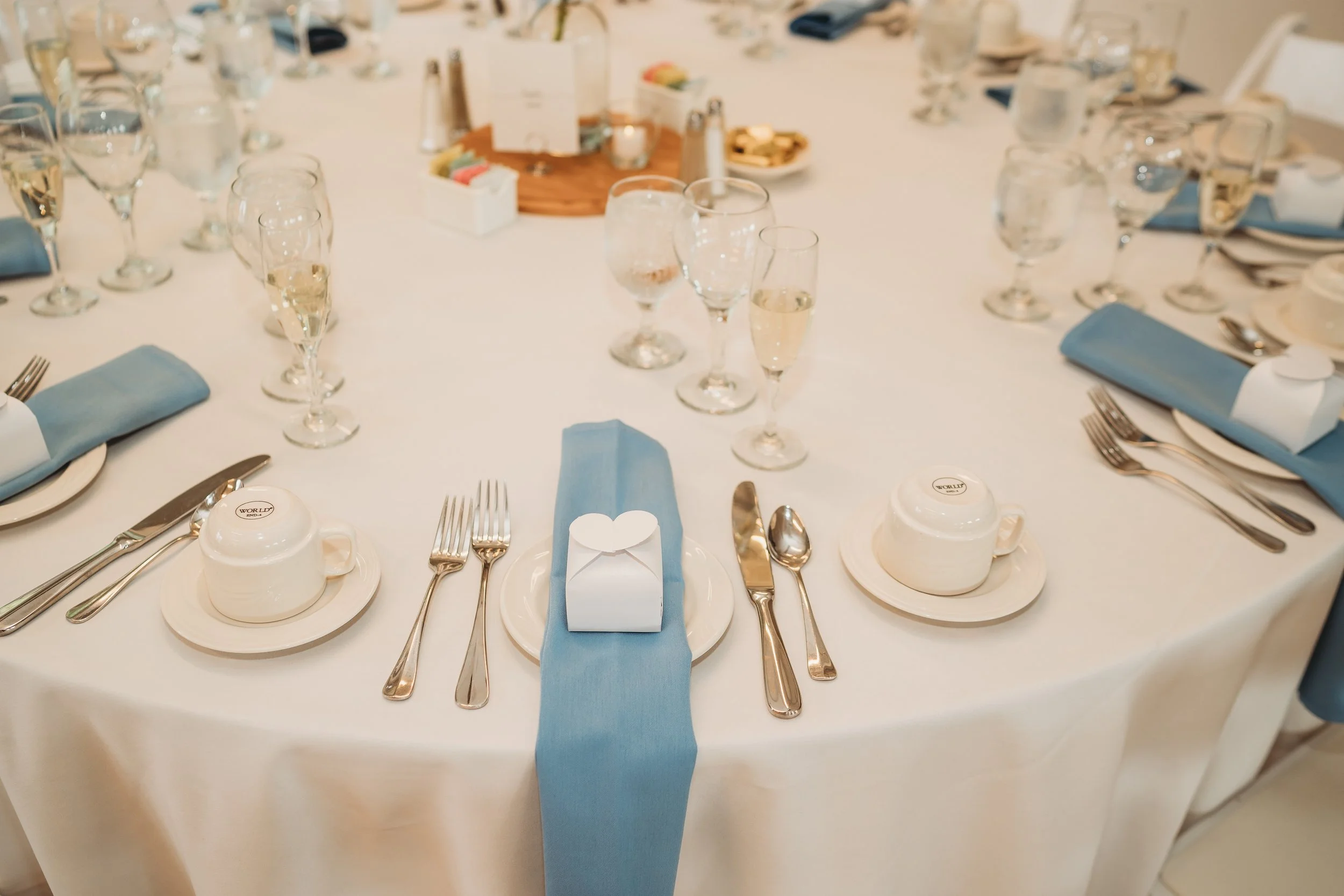 Formal dining table set with white tablecloth, blue napkins, white plates, cups, silverware, champagne glasses with bubbly, water glasses, and a centerpiece with condiments and a wooden platter.