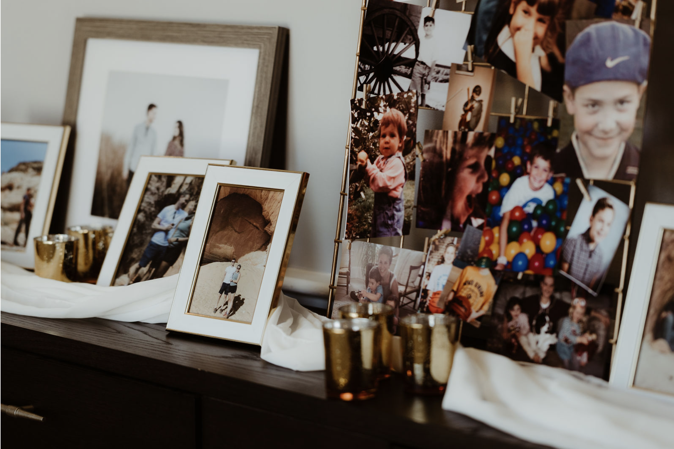 A display of framed photographs and a collage of printed photos on a table, with small gold-colored candle holders, set against a plain wall.