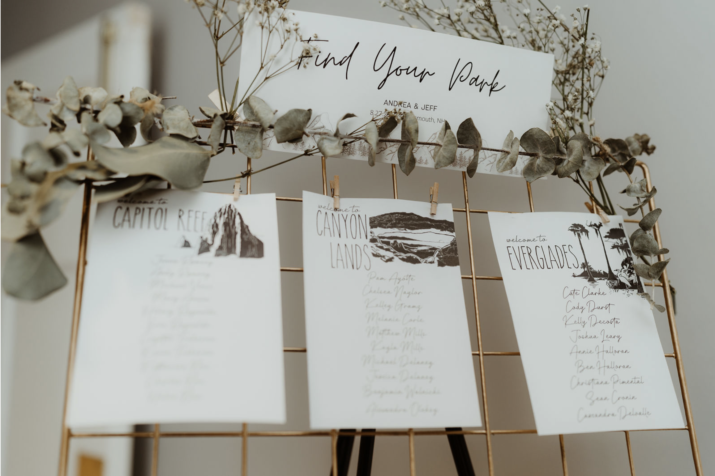 Welcome sign reading 'Find Your Path' displayed on a gold wire grid with dried eucalyptus leaves, with three printed list cards attached with small clothespins, each listing different locations and names for a wedding or event.