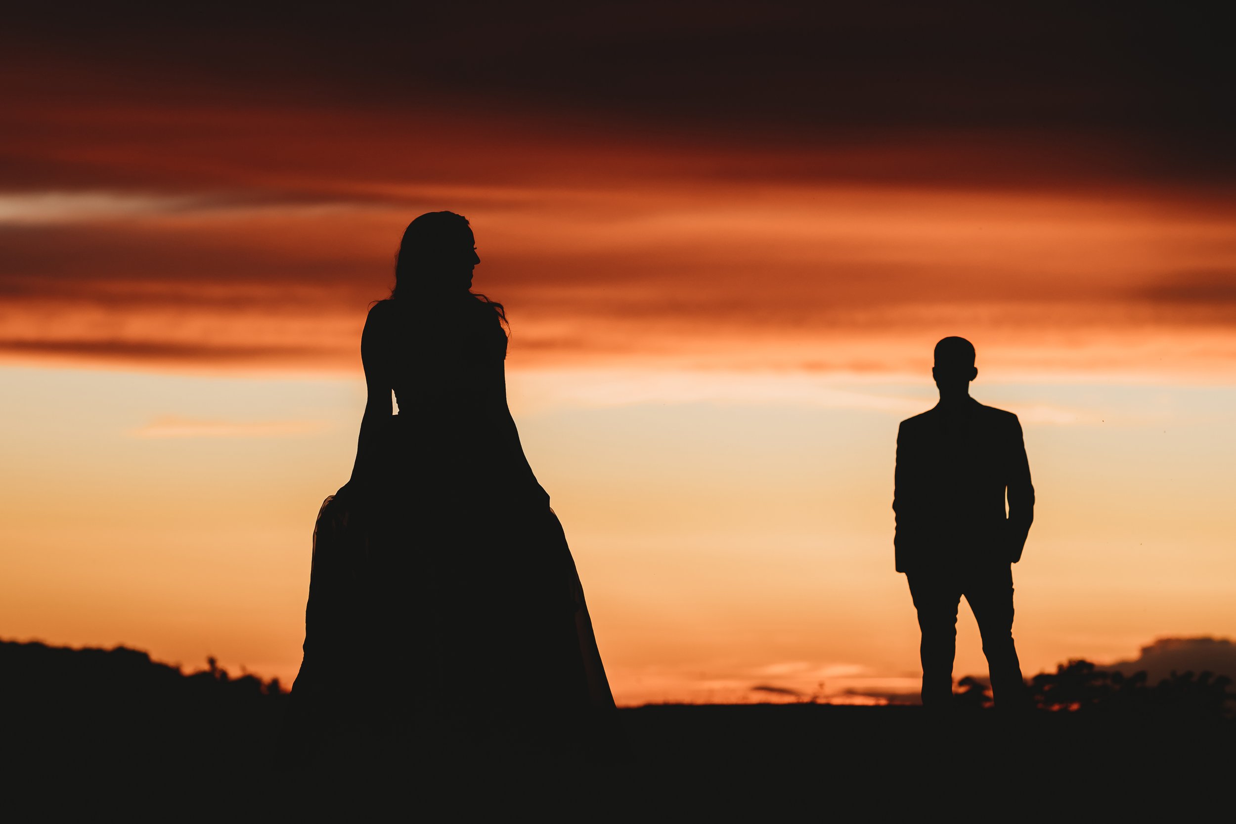 Silhouettes of a woman and a man standing apart during a sunset with a colorful sky in the background.
