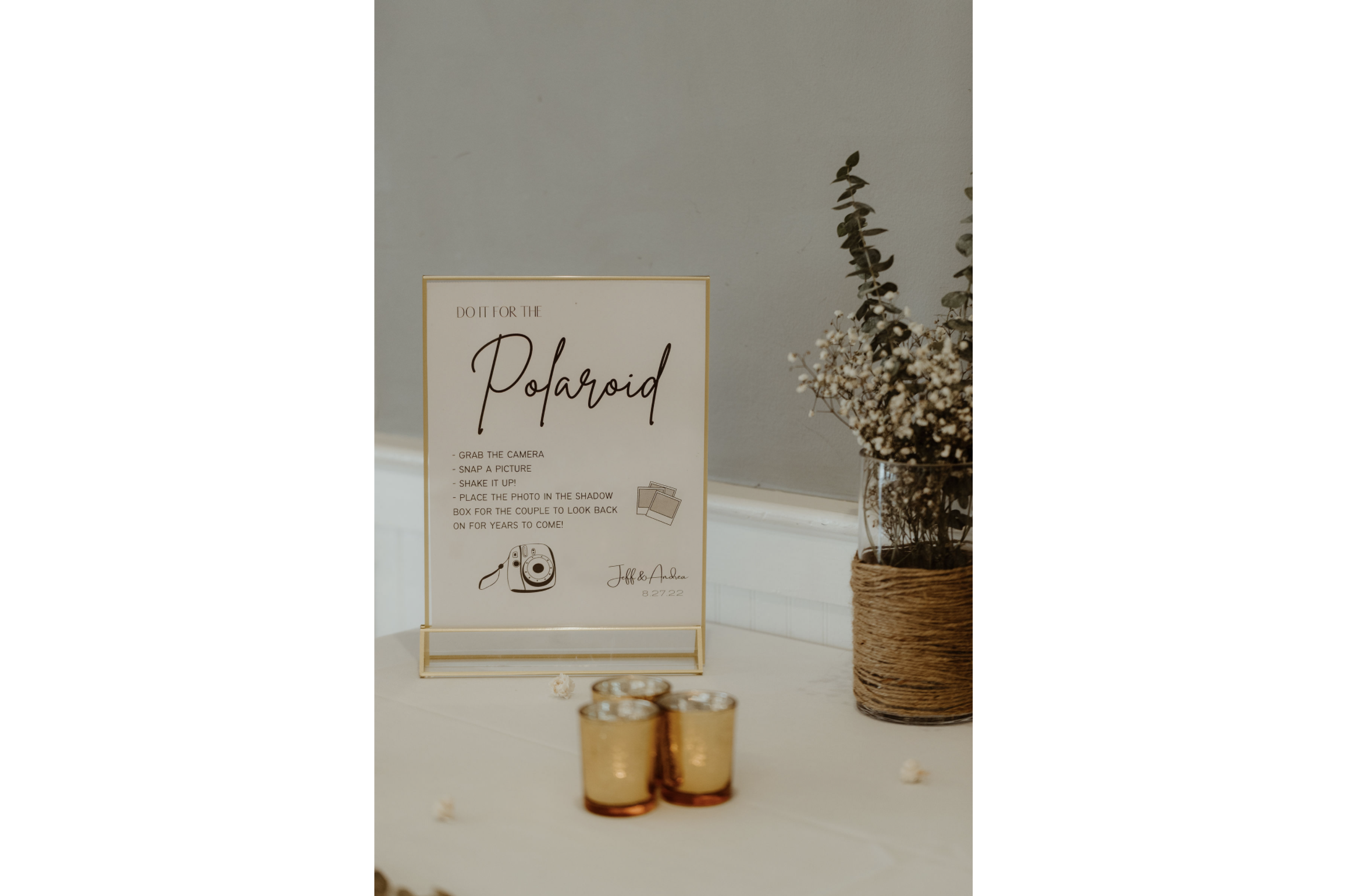A sign on a table with instructions for taking Polaroid photos, next to a glass vase filled with dried white flowers wrapped with twine, and three small gold candles.