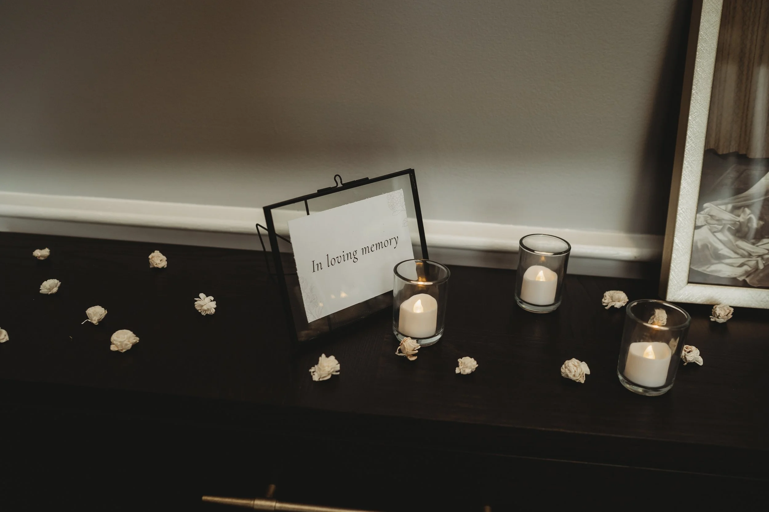 Candles in glass holders, scattered white flower petals, a framed memorial sign reading 'In loving memory', and a framed photograph.