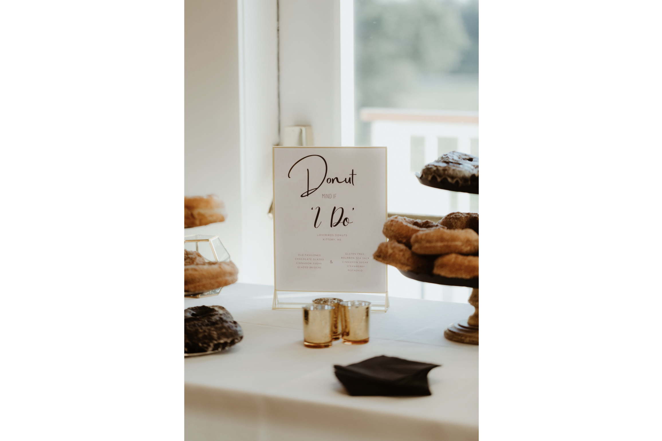 Table with assorted donuts on display, signs, and black napkins in a bright room with a window in the background.