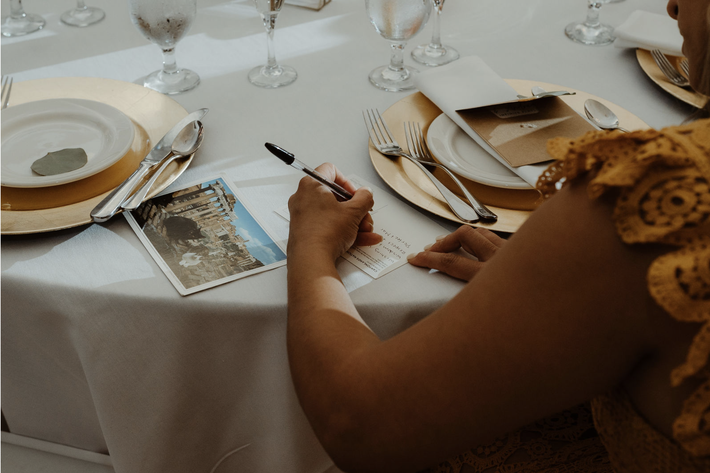 Person writing at a formal dining table with place settings, wine glasses, and a postcard of a cityscape.