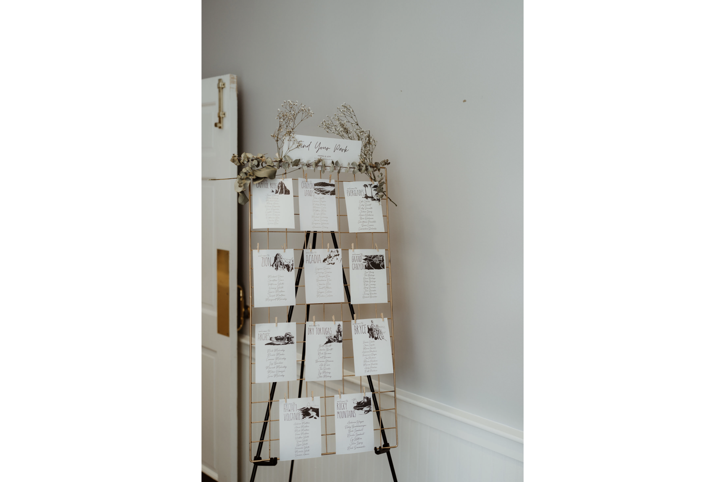 Guest seating chart display with illustrations of mountains and landscapes, decorated with dried flowers on a gold wire stand in a white-walled room.