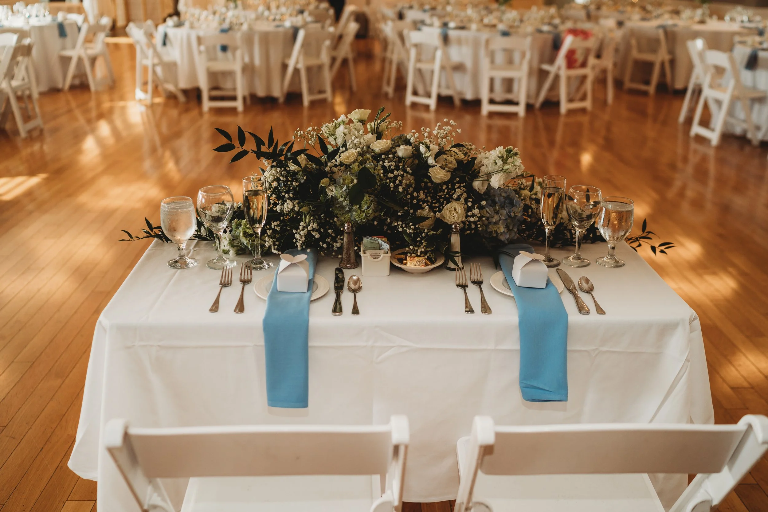 A banquet table decorated with a large floral centerpiece, surrounded by white chairs, with water glasses, plates, silverware, and blue napkins on a white tablecloth in a banquet hall.