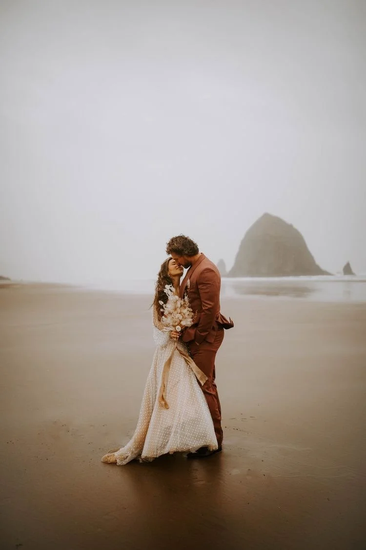 A couple dressed in wedding attire standing on a beach, embracing each other with a large rock formation in the background.