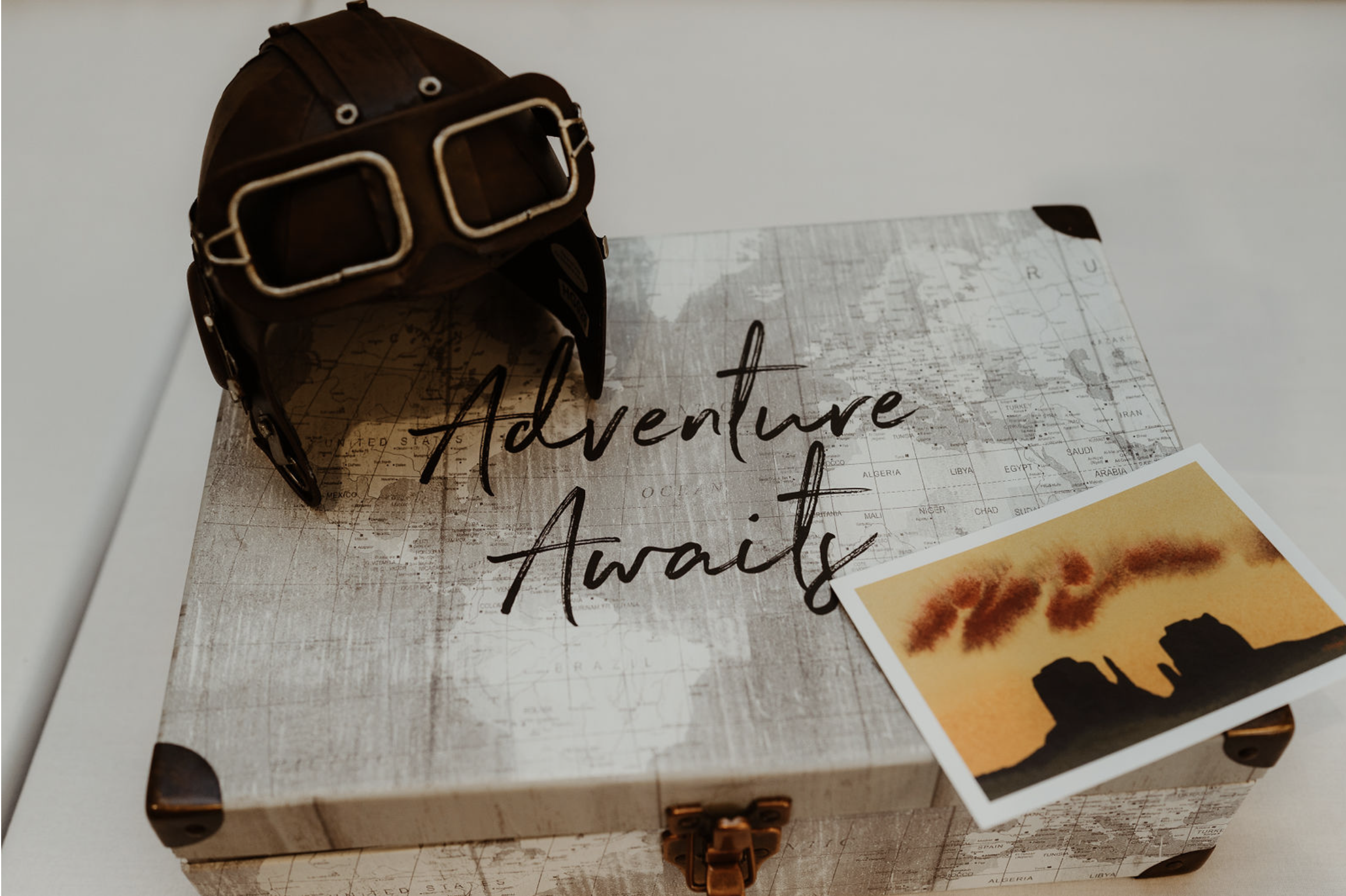 A vintage leather aviator helmet and goggles placed on a box with a map design, and a small photograph of a desert landscape with rock formations under an orange sky, with the words 'Adventure Awaits' written on the box.