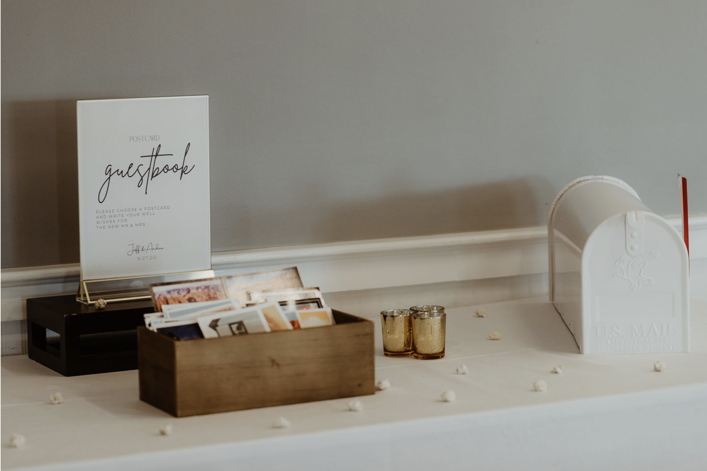 A wedding guestbook display on a table with a sign, a wooden box filled with postcards, two small candles, and a white mailbox for guest messages.