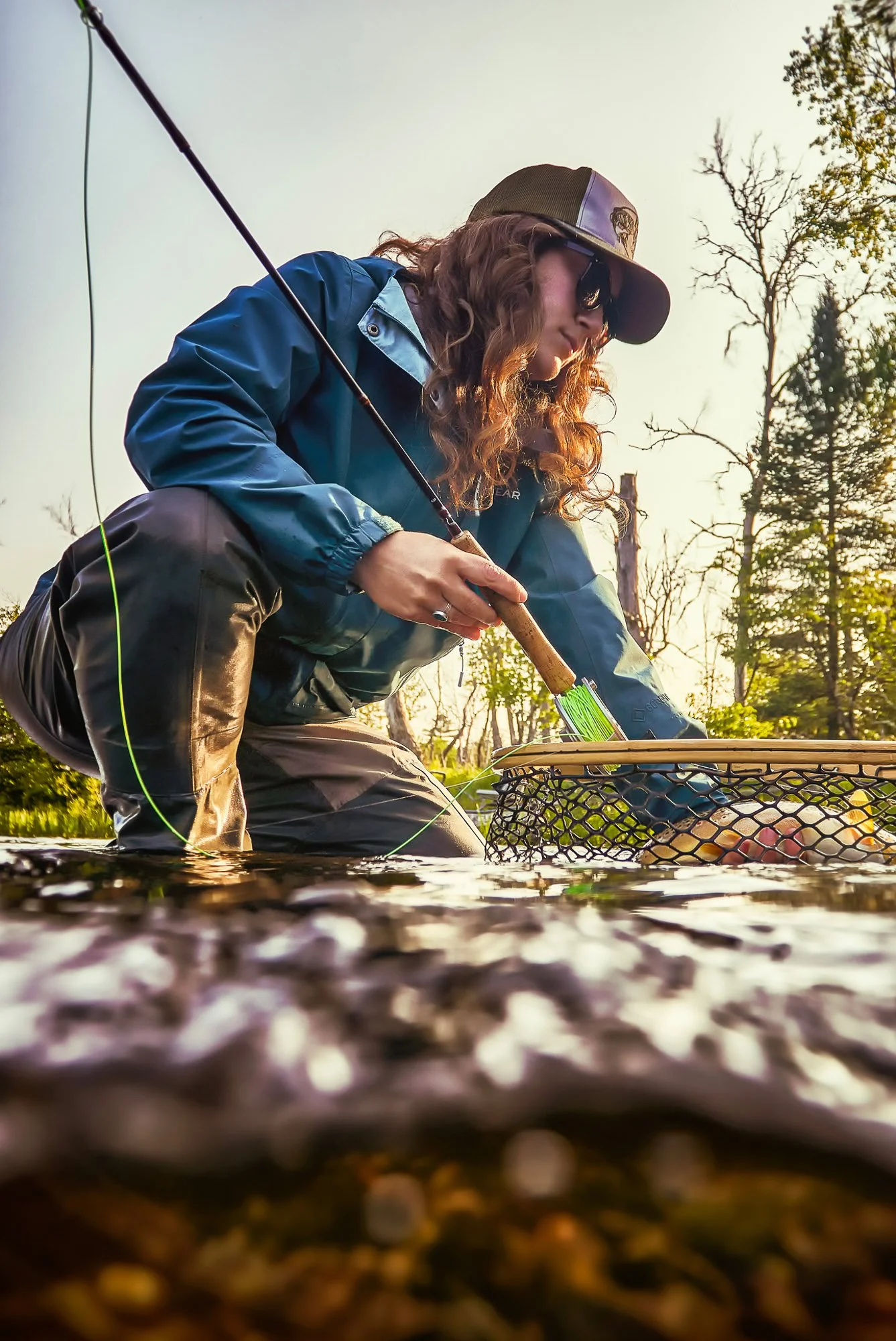 Person in outdoor clothing, fishing rod, and sunglasses kneeling in a river, handling a fish in a basket during sunset with trees in the background.