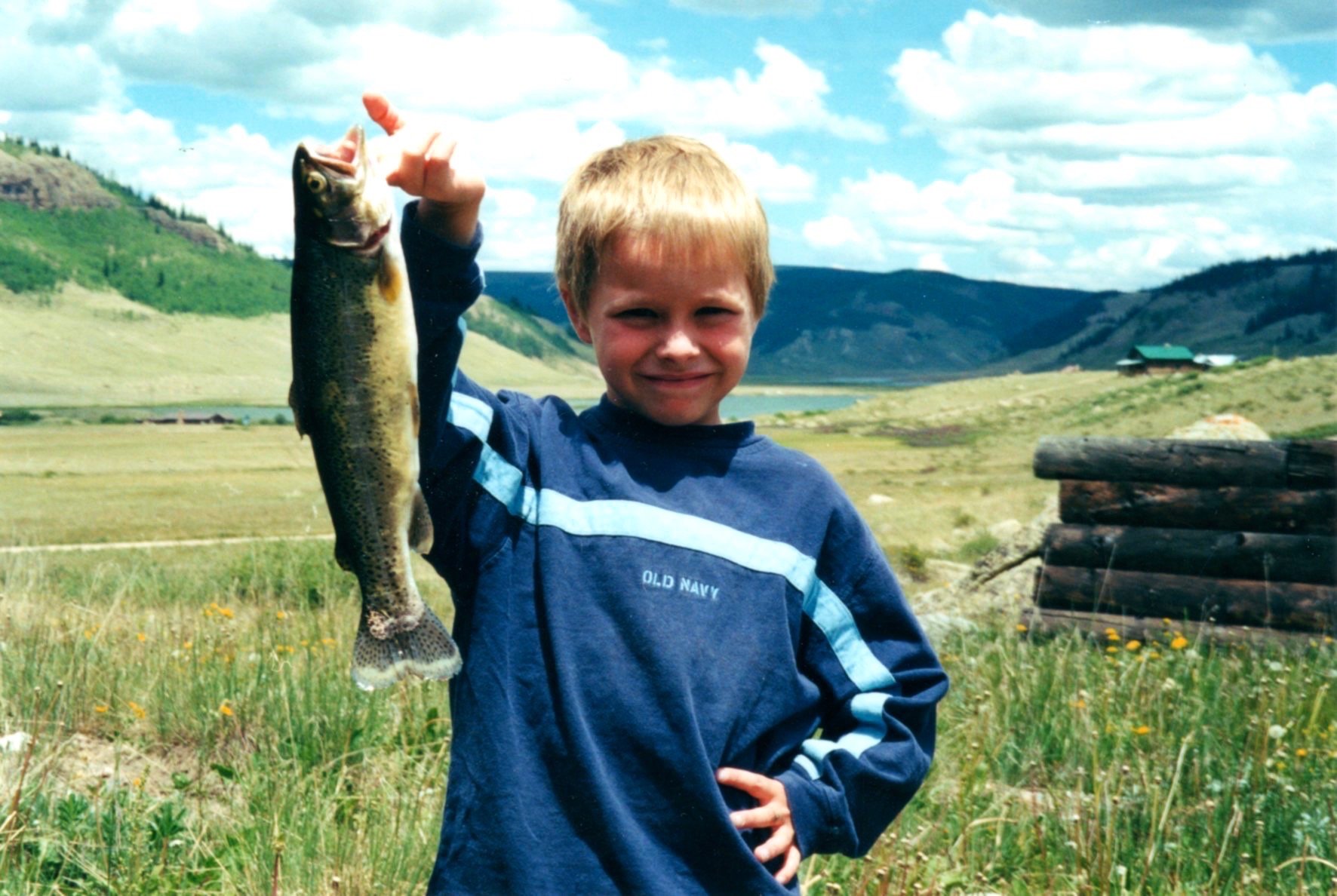 A young boy with blonde hair and a blue sweatshirt, smiling and holding a fish in one hand, standing outdoors in a grassy field with mountains and a partly cloudy sky in the background.