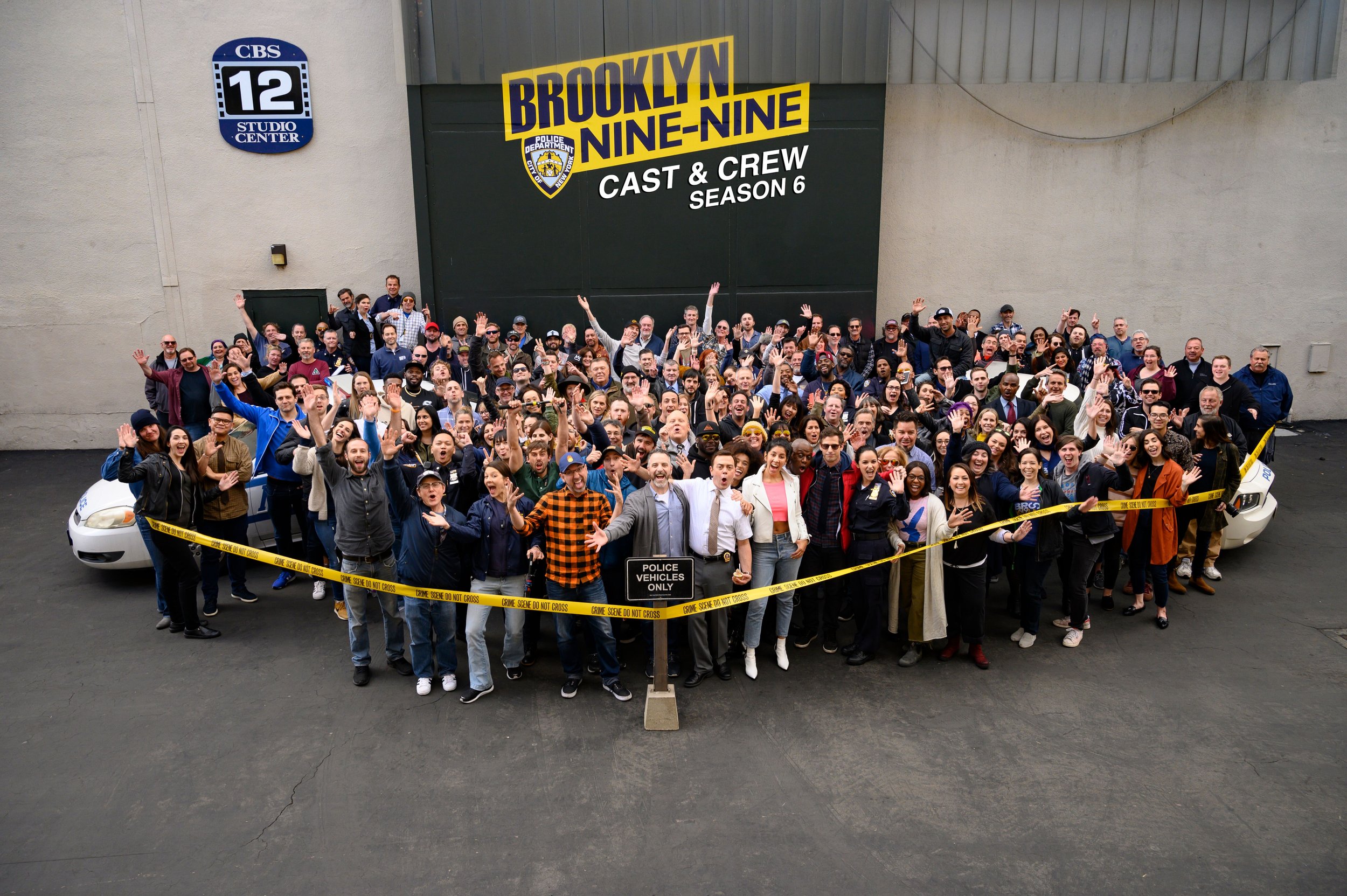 Large group of people posing together in front of a building with a sign that reads "Brooklyn Nine-Nine" and "Cast & Crew Season 6," surrounded by yellow police crime scene tape.
