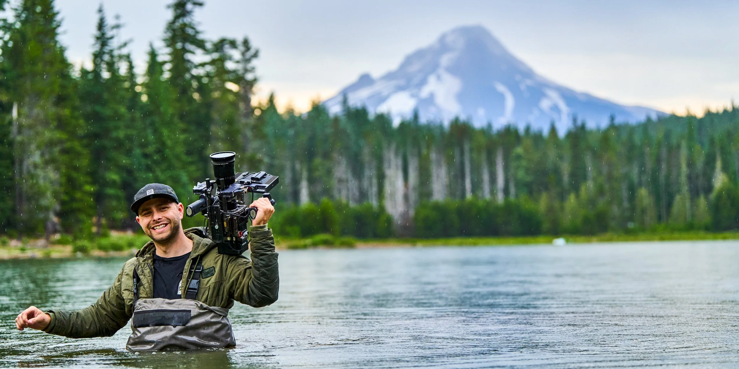 A man standing in a river holding a professional video camera on his shoulder, with a forest and snow-capped mountain in the background.